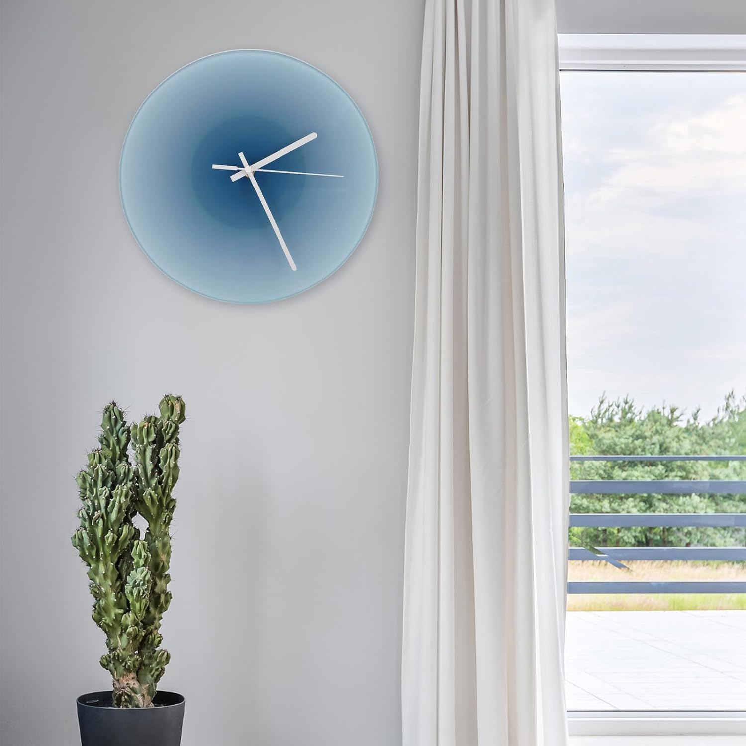 Blaue Wanduhr hängt an weißer Wand, links daneben ein grüner Kaktus im Topf, rechts ein Fenster mit Vorhang.