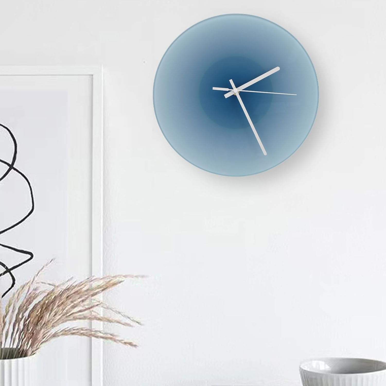 Eine runde blaue Wanduhr mit weißen Zeigern auf einer weißen Wand neben gerahmter Kunst und getrocknetem Gras.