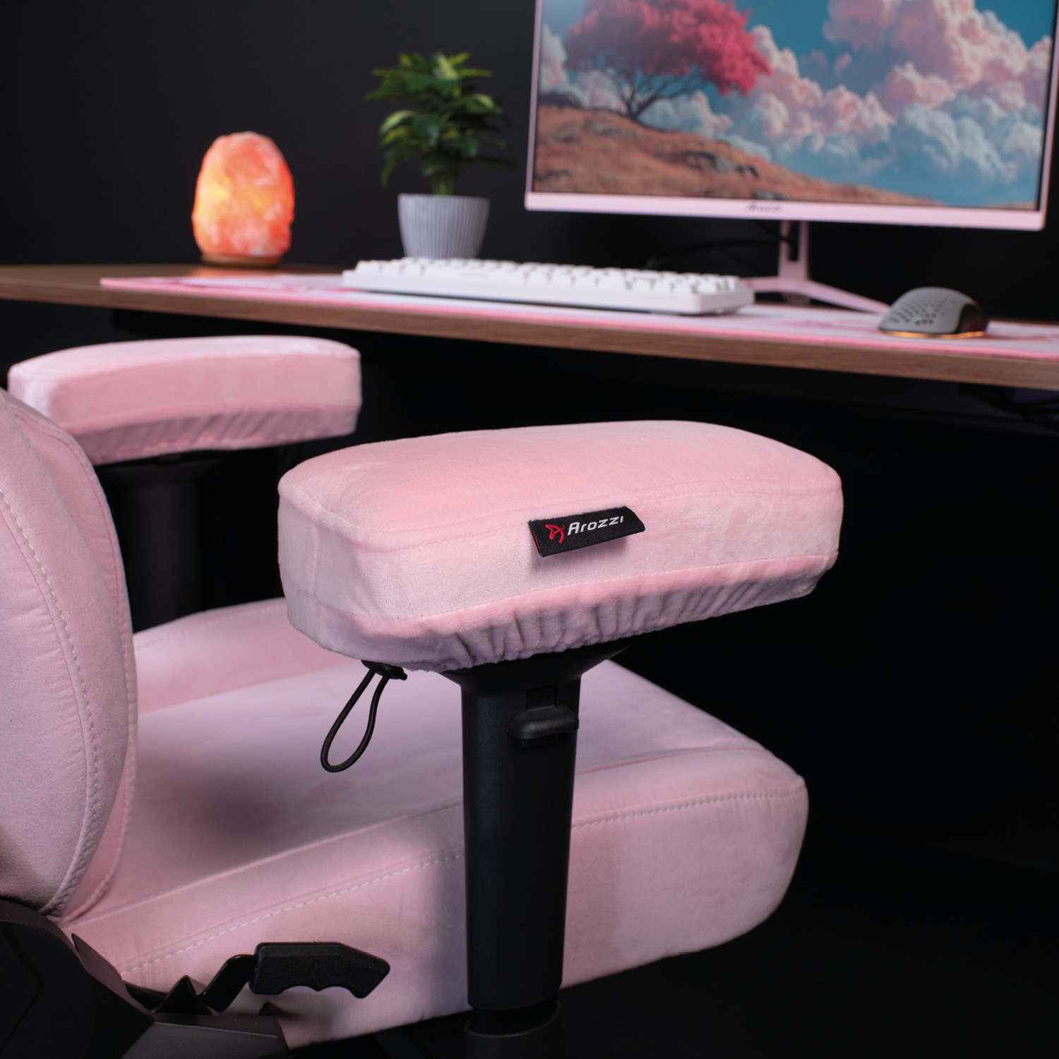 Una sedia da gaming rosa con braccioli in peluche in primo piano, con scrivania, tastiera, monitor e lampada di sale sullo sfondo.