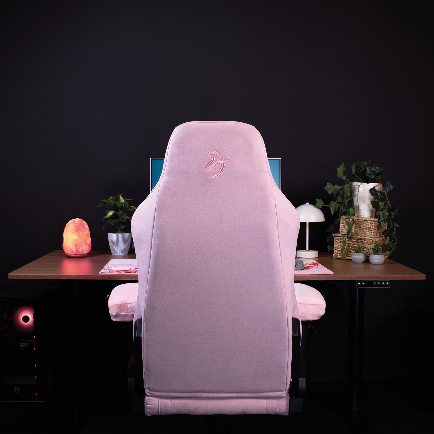 Una sedia da gaming rosa davanti a una scrivania in legno con PC, lampada, piante e lampada di sale su sfondo nero.