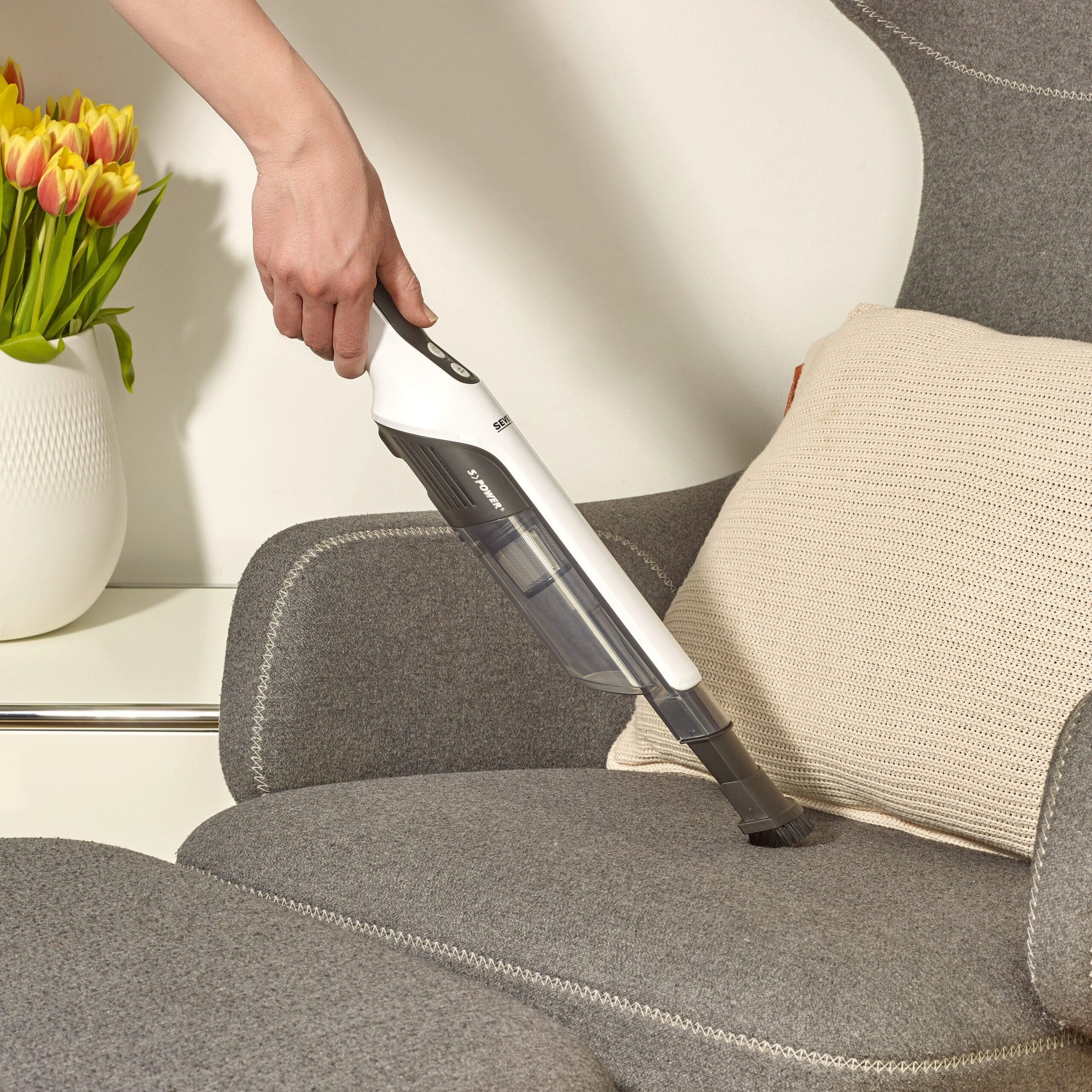 Une main aspire un fauteuil gris avec un aspirateur à main blanc. Un vase avec des tulipes jaunes est à l'arrière-plan.