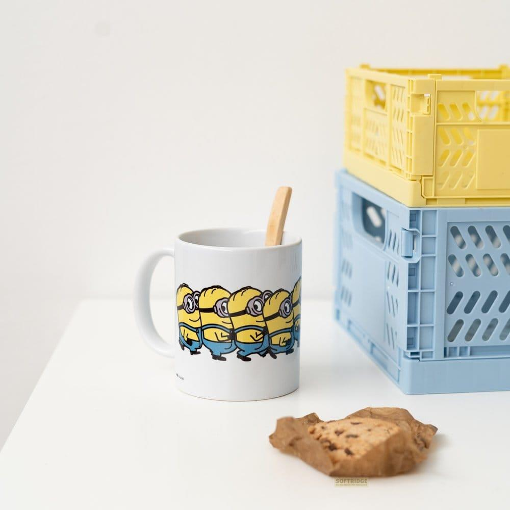 Una taza blanca con Minions, una cuchara de madera, una galleta en papel y cajas de plástico apiladas amarilla y azul en una mesa blanca.