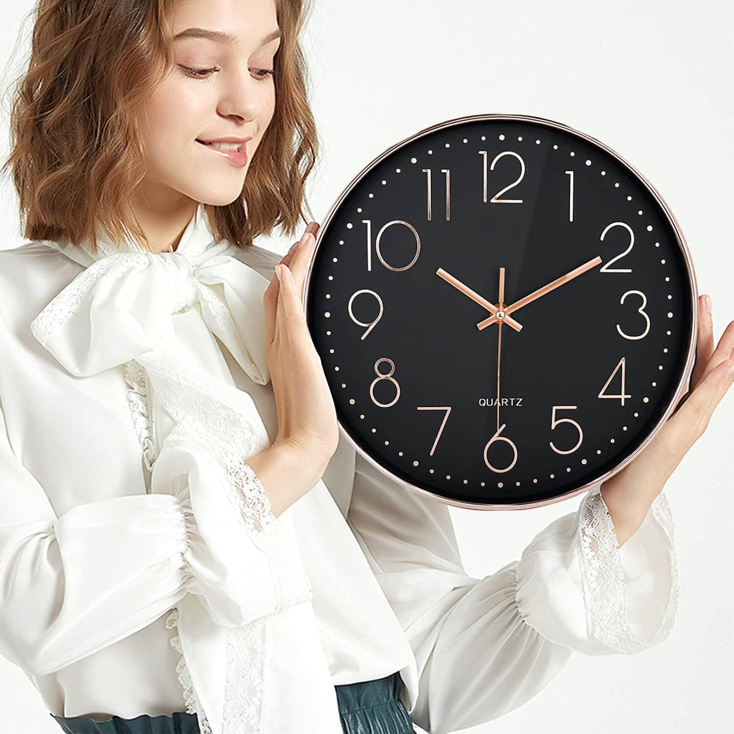 Eine Frau in weißer Bluse hält eine große, schwarze Wanduhr mit roségoldenen Zahlen und Zeigern.