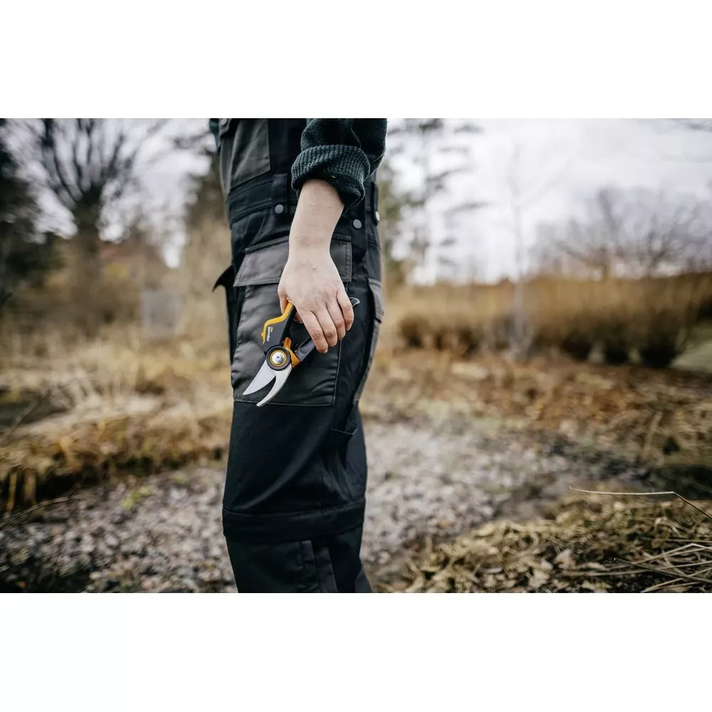 Person in Arbeitskleidung hält eine Gartenschere in der Hand, im Hintergrund ein Garten.