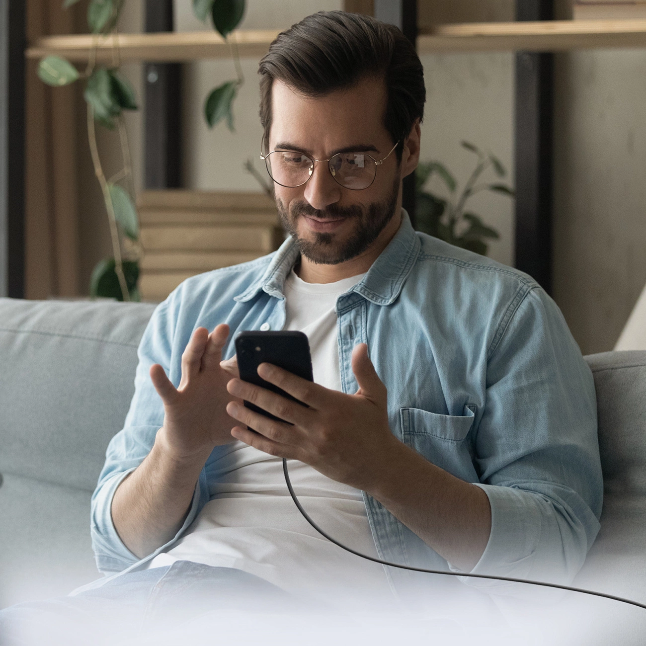 Un hombre con barba, gafas y camisa vaquera está sentado en un sofá usando un smartphone.