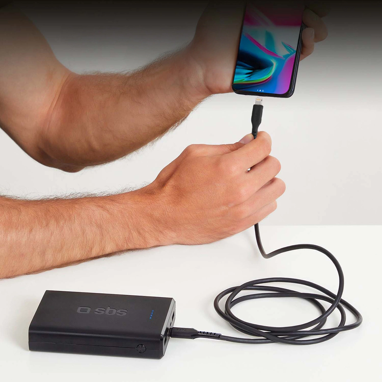 Una persona collega uno smartphone a un power bank SBS nero con un cavo di ricarica su sfondo bianco.