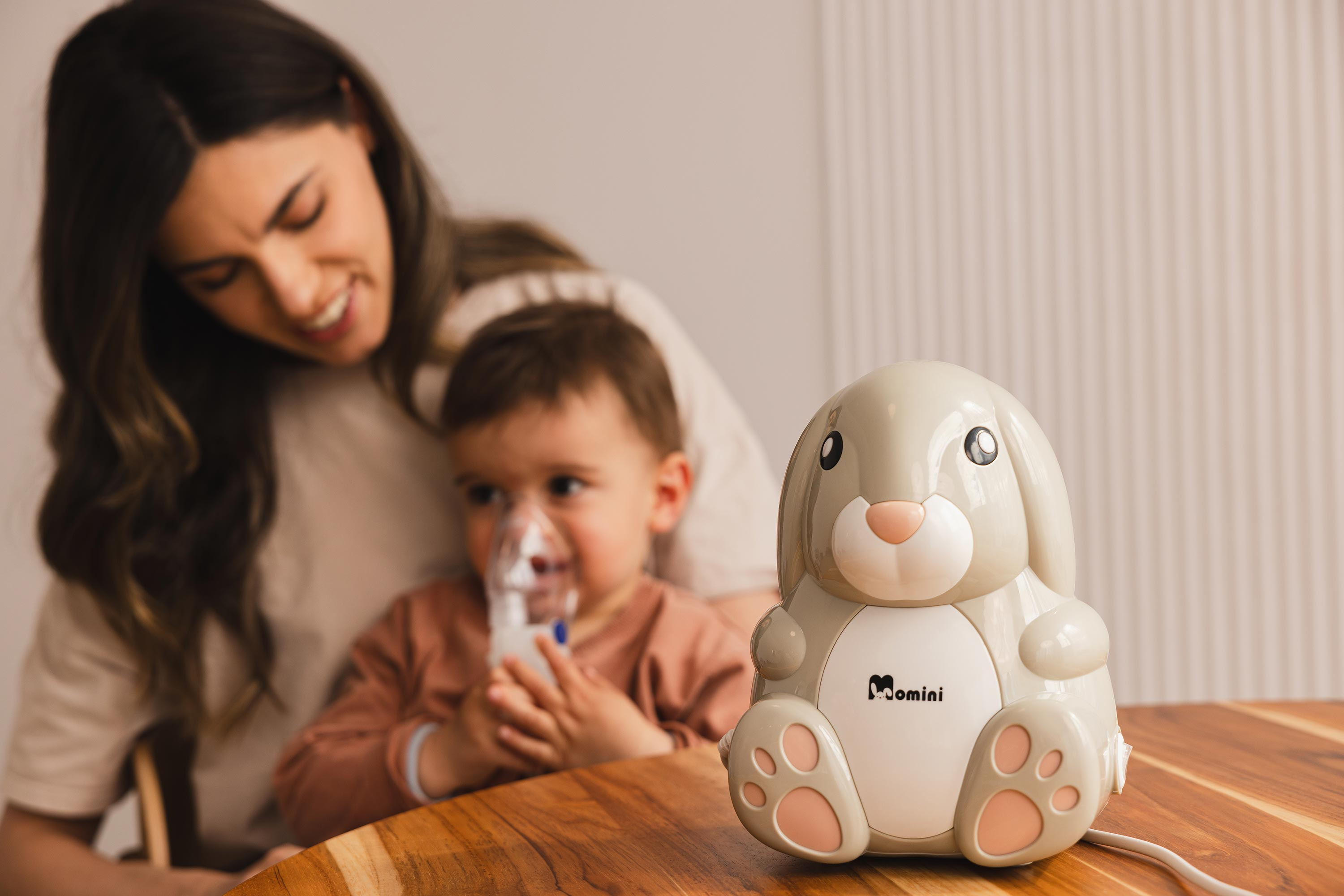 Ein grauer, kaninchenförmiger Inhalator steht auf einem Holztisch, im verschwommenen Hintergrund nutzen Mutter und Kind ein Gerät.
