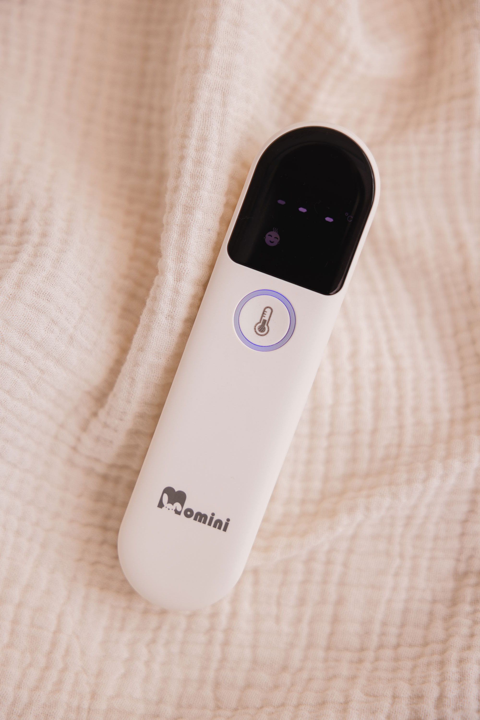 Ein weißes Momini-Infrarot-Thermometer mit digitalem Display liegt auf einer beigen Musselindecke.