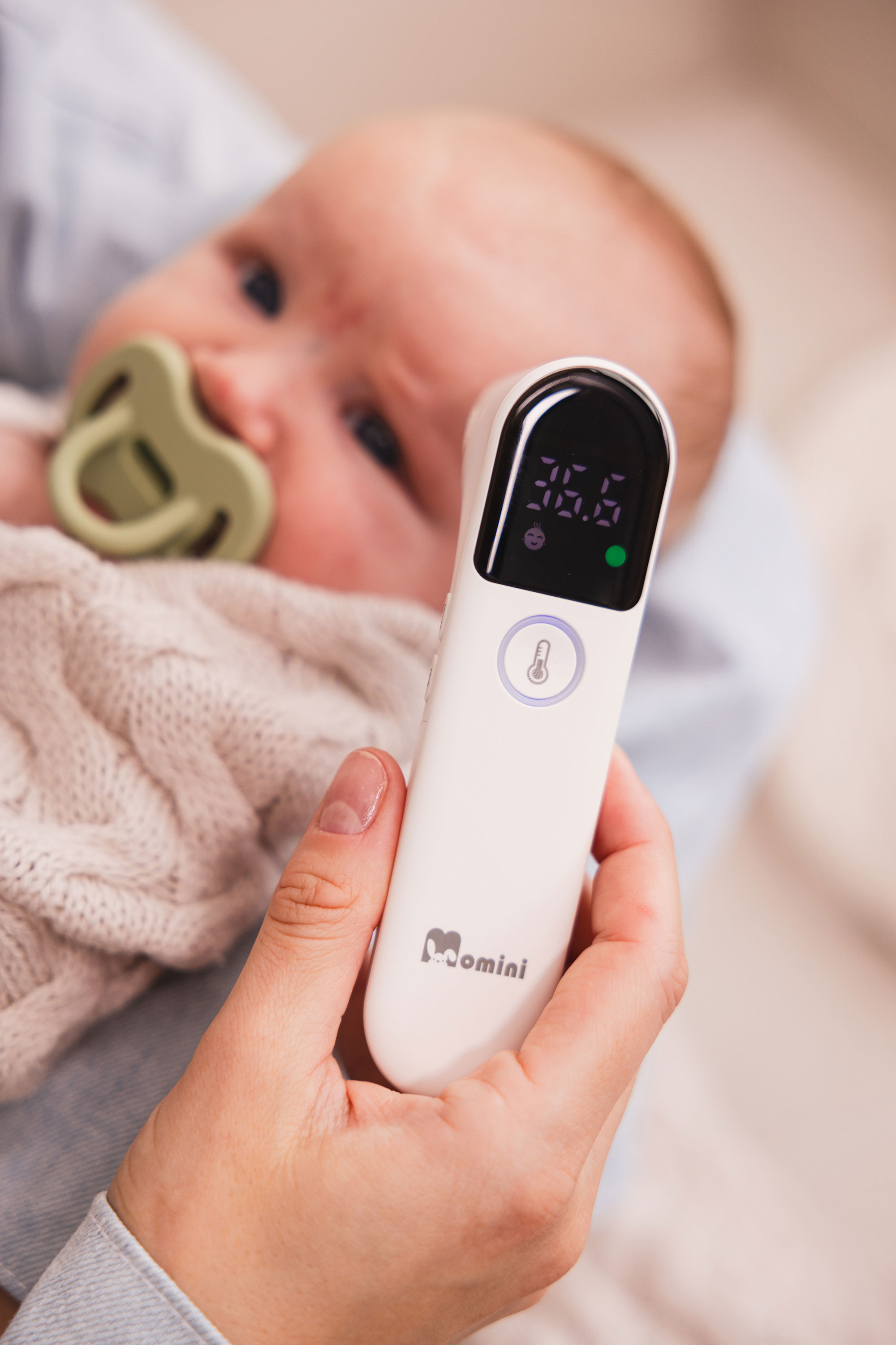 Eine Hand hält ein weißes digitales Infrarot-Thermometer, im Hintergrund ein Baby mit Schnuller.