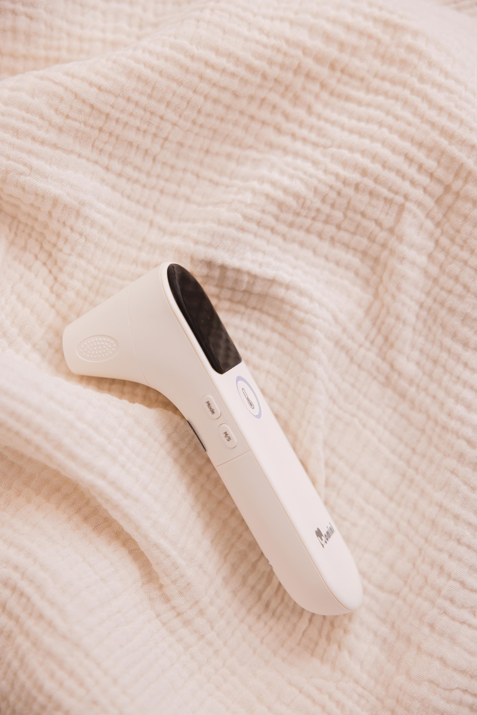 Ein weißes Infrarot-Thermometer liegt auf einer beigen, faltigen Stoffdecke.