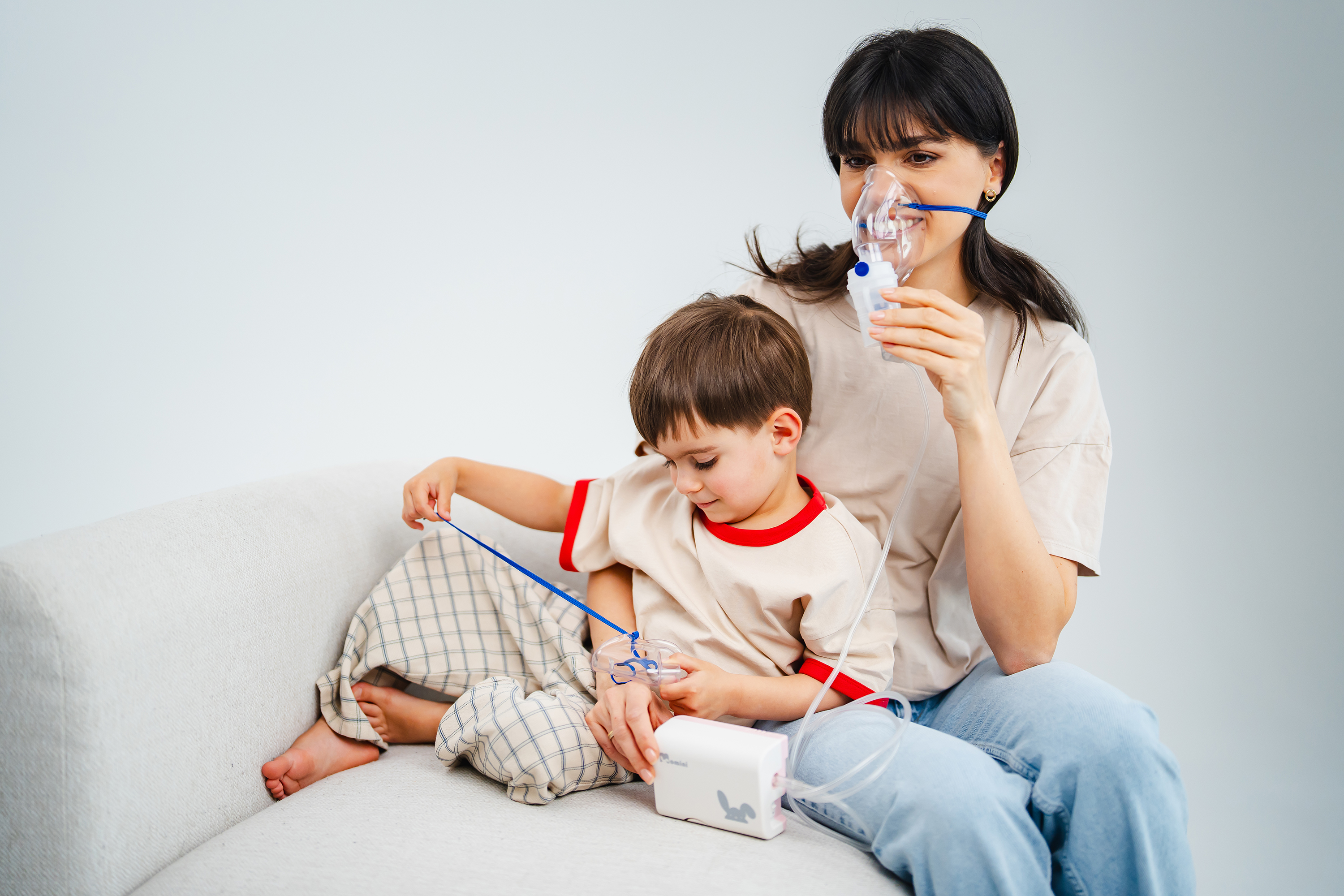 Eine Mutter und ihr Sohn sitzen auf einem Sofa und benutzen ein Inhalationsgerät mit Masken.