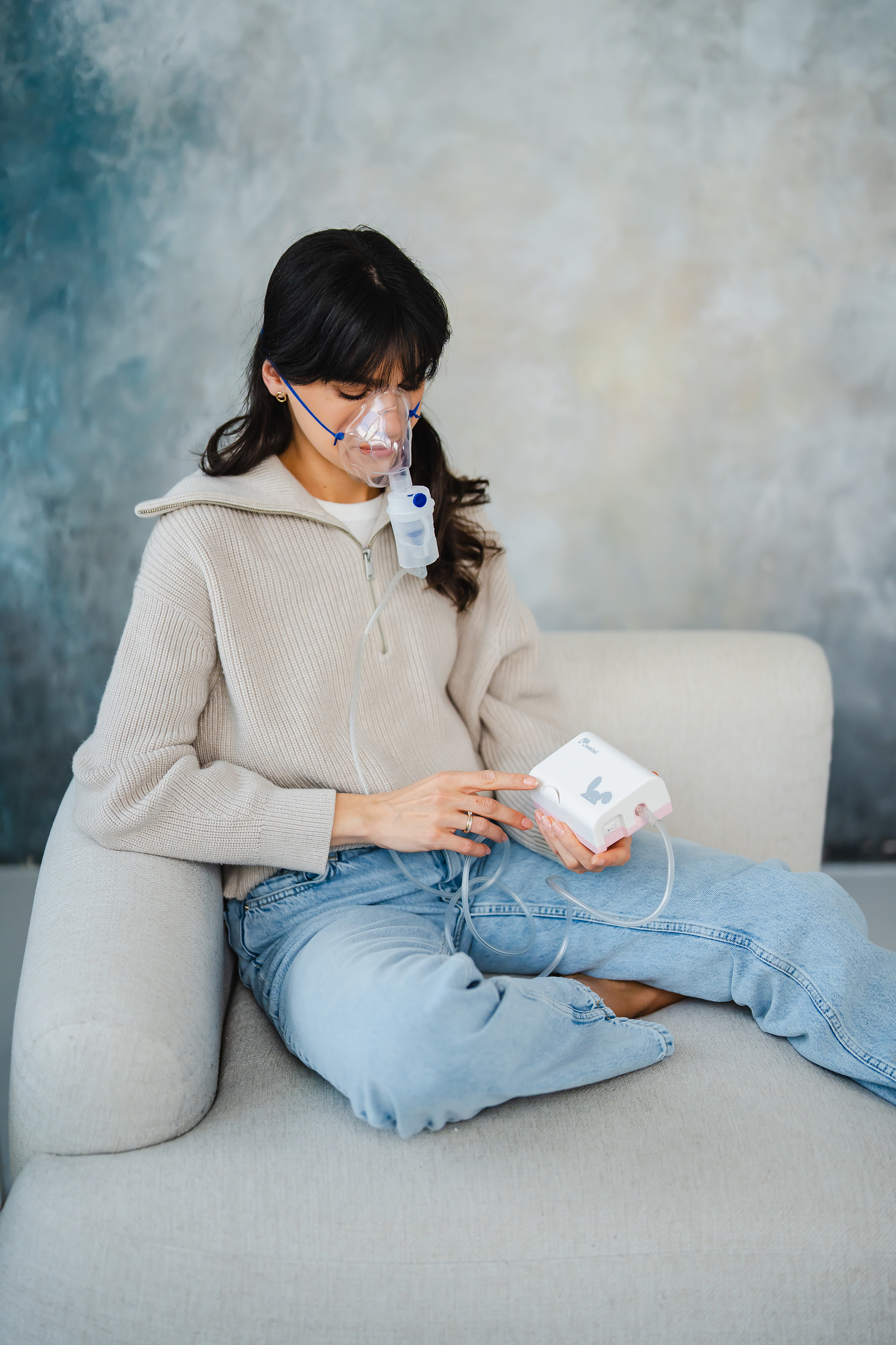 Eine Frau mit dunklen Haaren sitzt auf einem weißen Sofa, trägt eine Sauerstoffmaske und bedient ein Inhalationsgerät.
