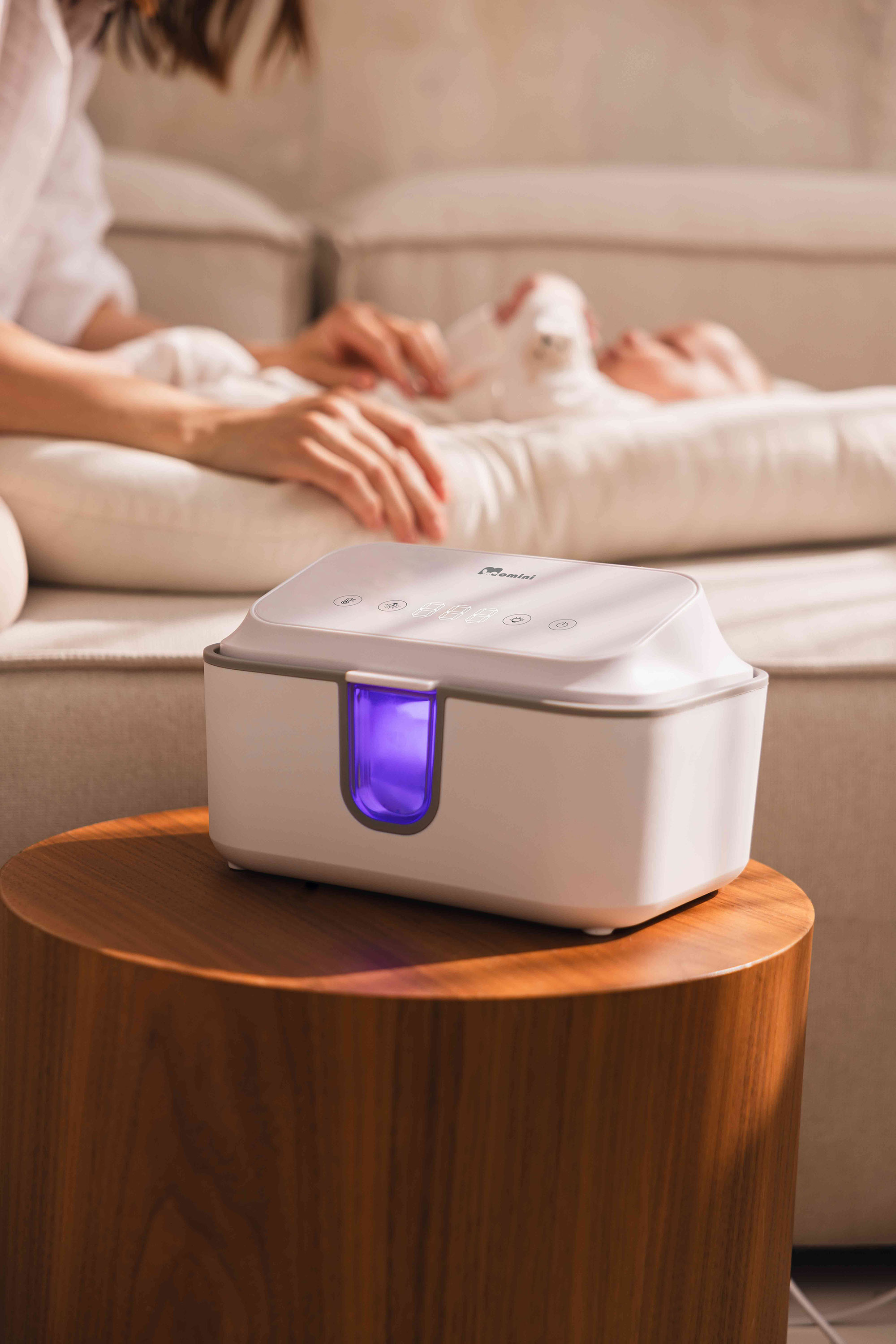 Biały sterylizator UV z fioletowym światłem na drewnianym stoliku, w tle kobieta z dzieckiem.