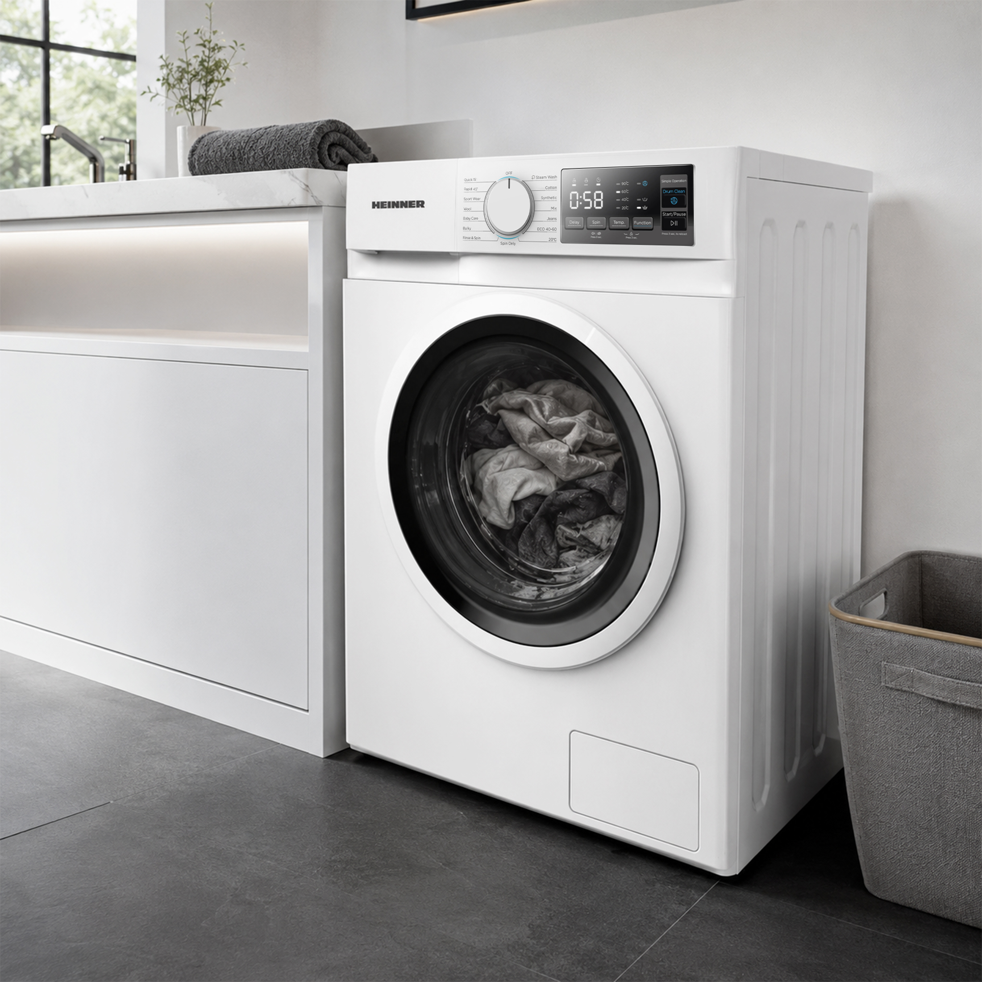 Un lave-linge blanc Heinner dans une salle de bain moderne, rempli de linge, à côté d'un panier gris.