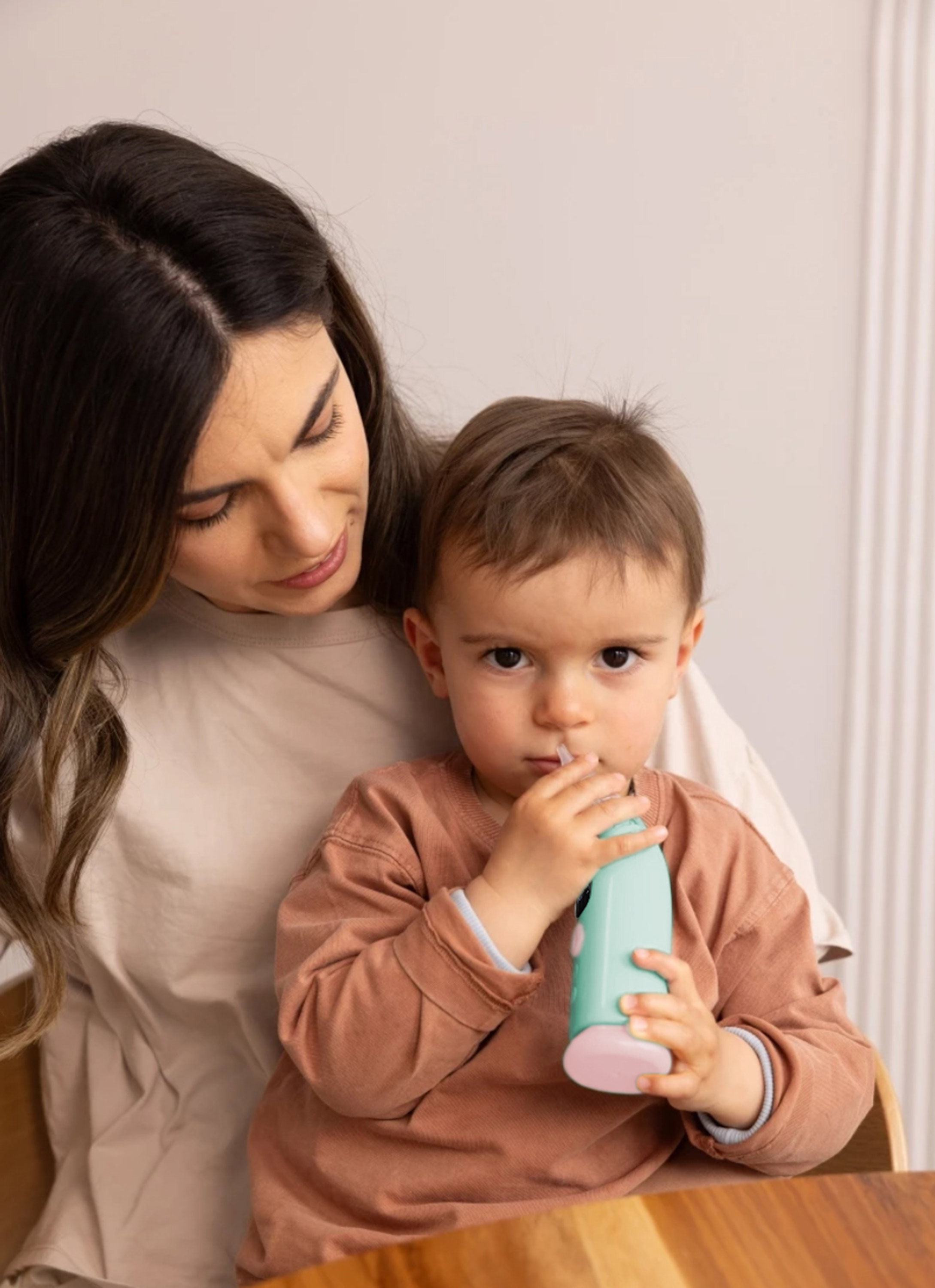 Una madre tiene in braccio il suo bambino, che usa un aspiratore nasale azzurro, seduto a un tavolo di legno.