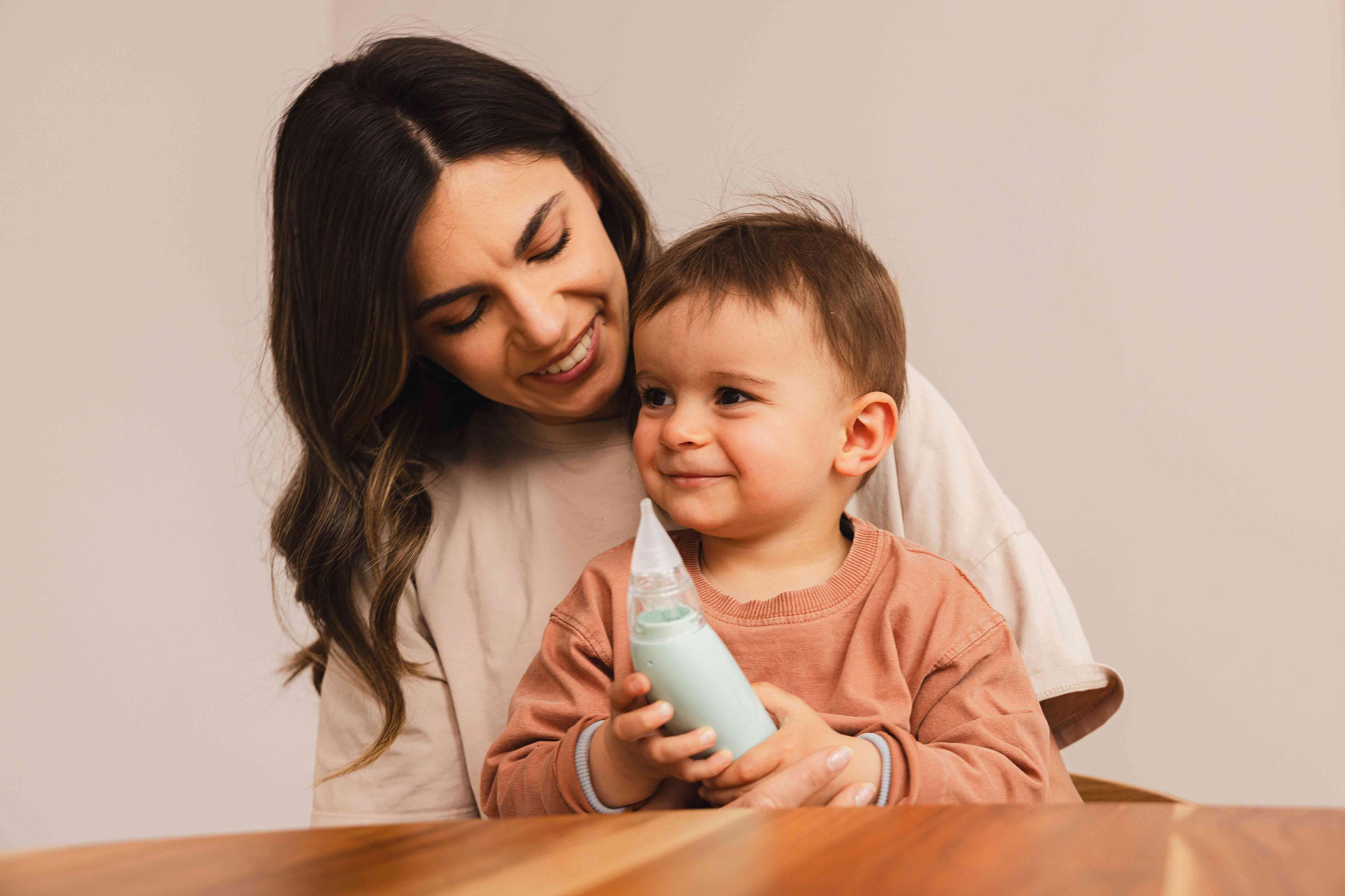 Una madre sorridente tiene in braccio il suo bambino che tiene un aspiratore nasale verde.