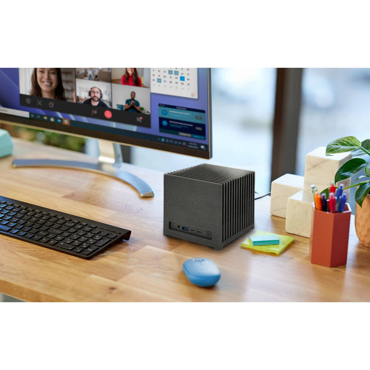 Un mini PC su una scrivania di legno vicino a tastiera, mouse blu e un monitor con videoconferenza.