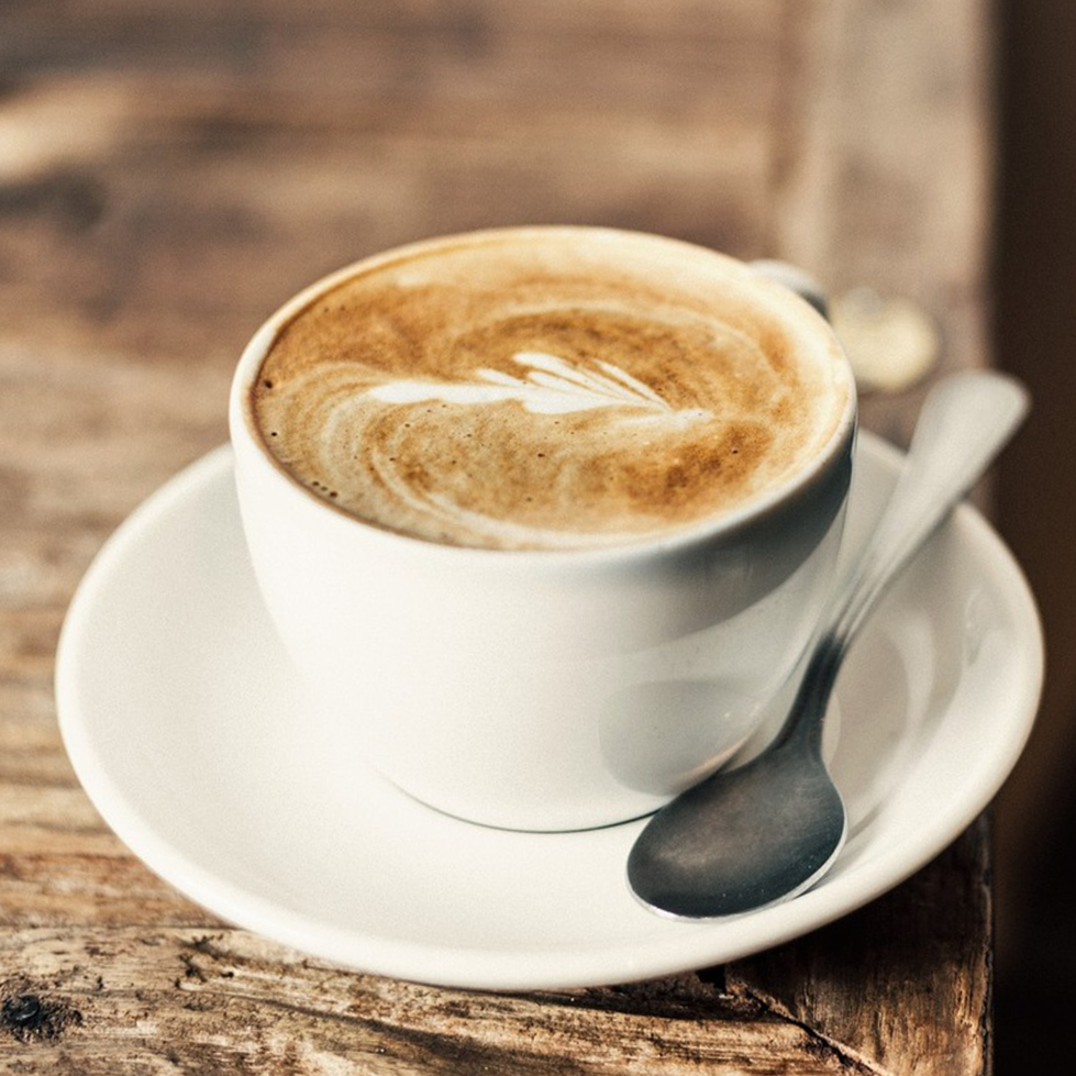 Biała filiżanka cappuccino z latte art na spodeczku z łyżeczką, na drewnie.