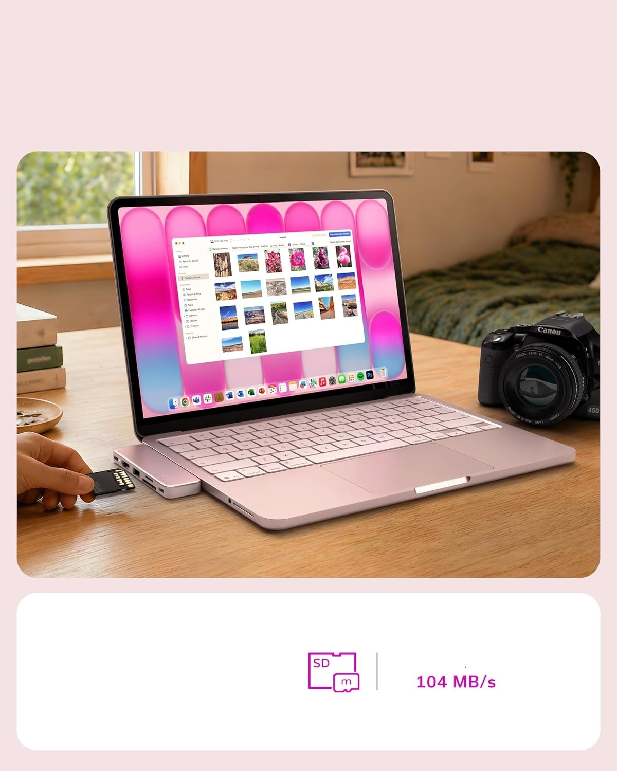 Różowy laptop na biurku, ręka wsadza kartę SD do huba; w tle aparat i książki.