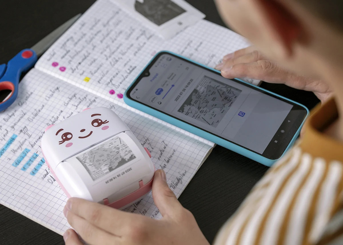 Un bambino usa uno smartphone per azionare una piccola e simpatica stampante fotografica su un quaderno a quadretti.