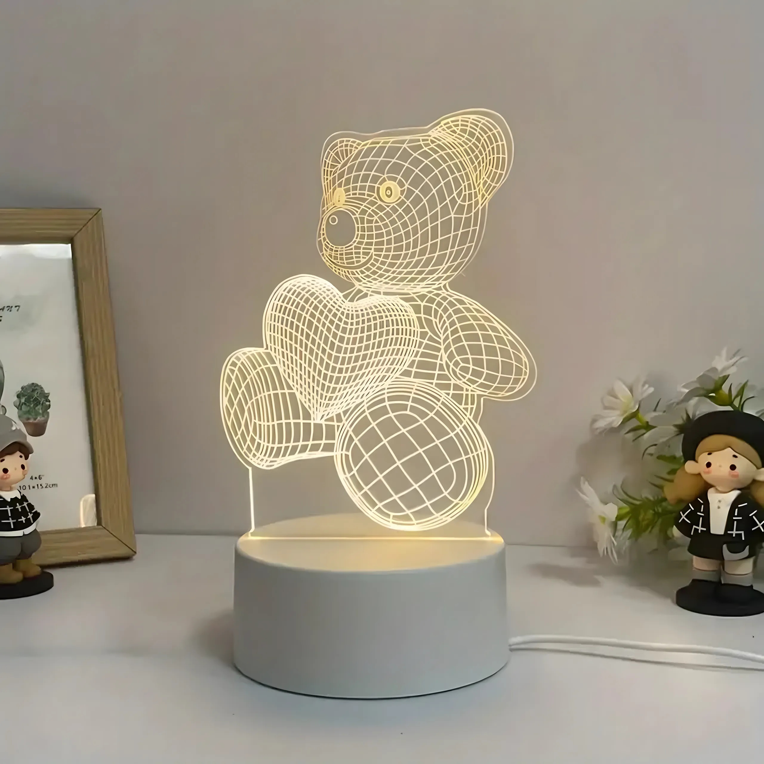 Podświetlana lampka 3D z misiem trzymającym serce na białej podstawie, w otoczeniu ramki na zdjęcia i figurki.