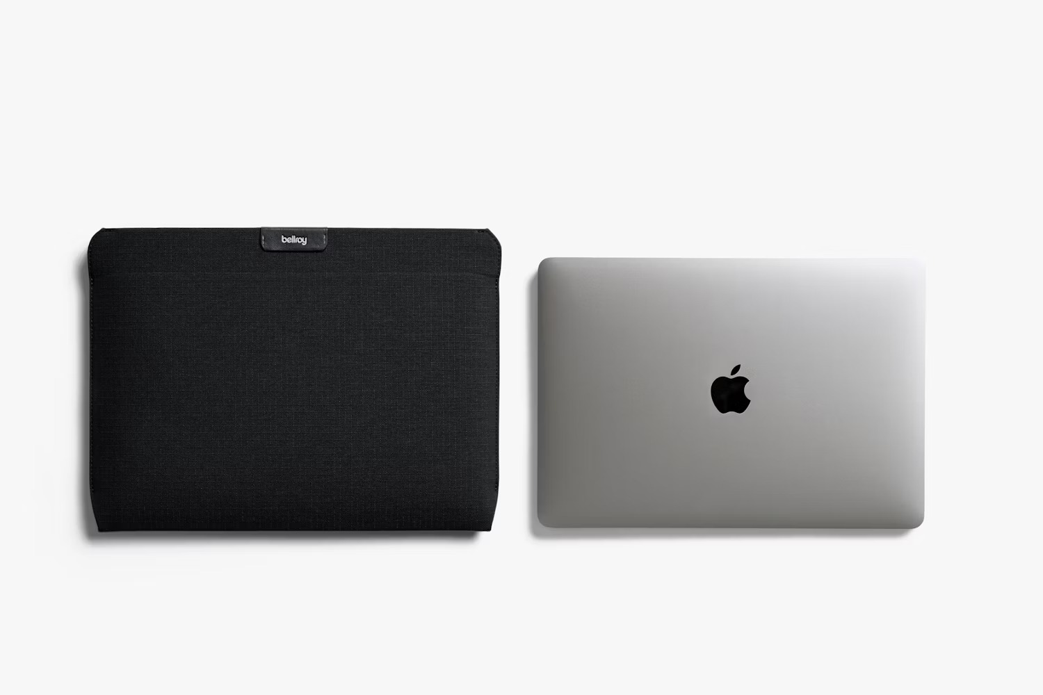 Czarny pokrowiec na laptopa Bellroy leży obok zamkniętego srebrnego MacBooka na białym tle.
