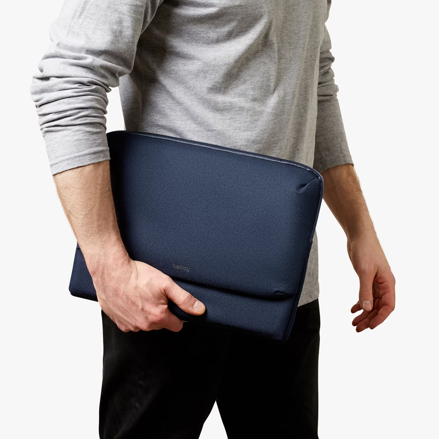 Osoba w szarej koszuli z długim rękawem i czarnych spodniach trzyma ciemnoniebieskie etui na laptopa na białym tle.