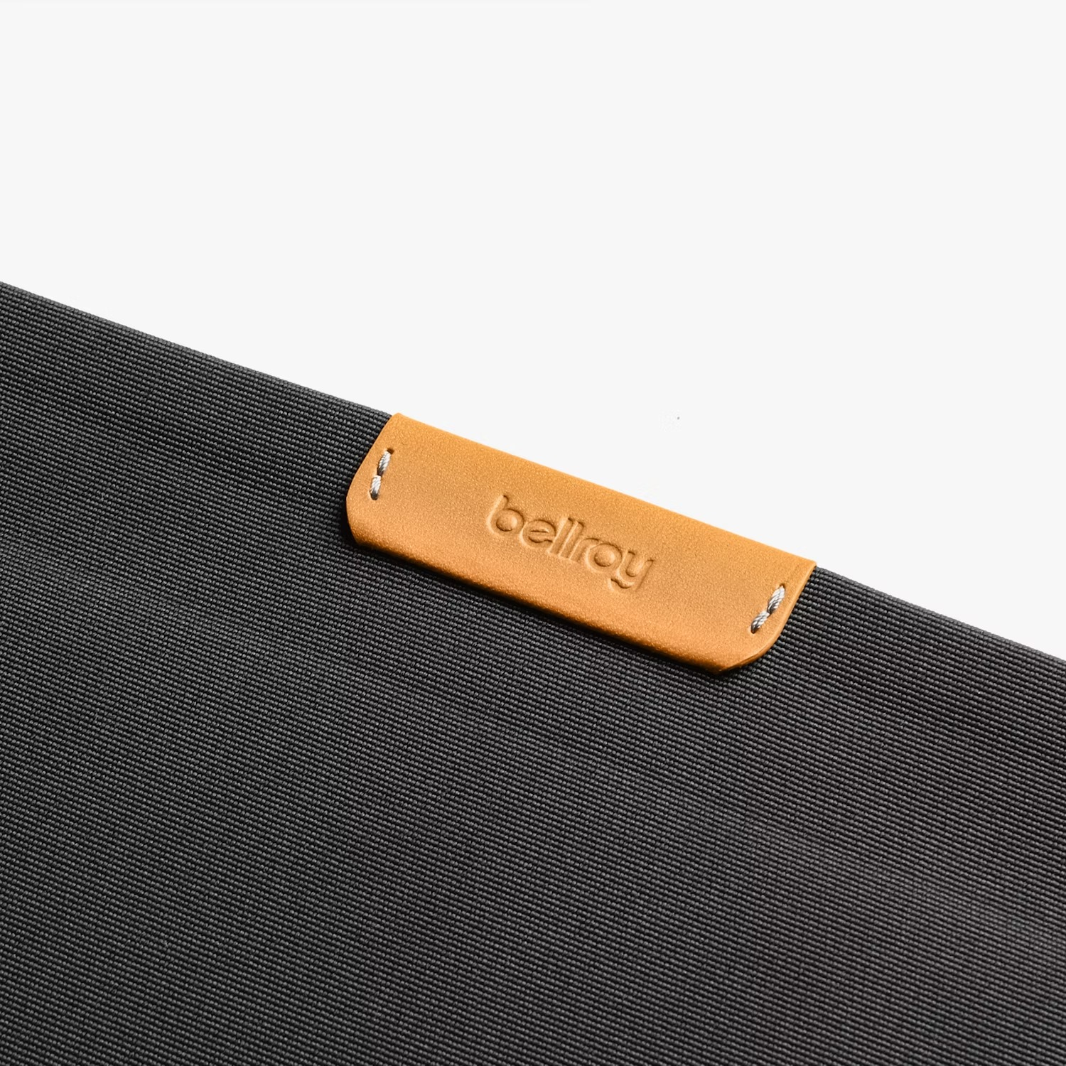 Zbliżenie ciemnoszarej powierzchni materiału z jasnobrązową skórzaną metką i wytłoczonym logo Bellroy.
