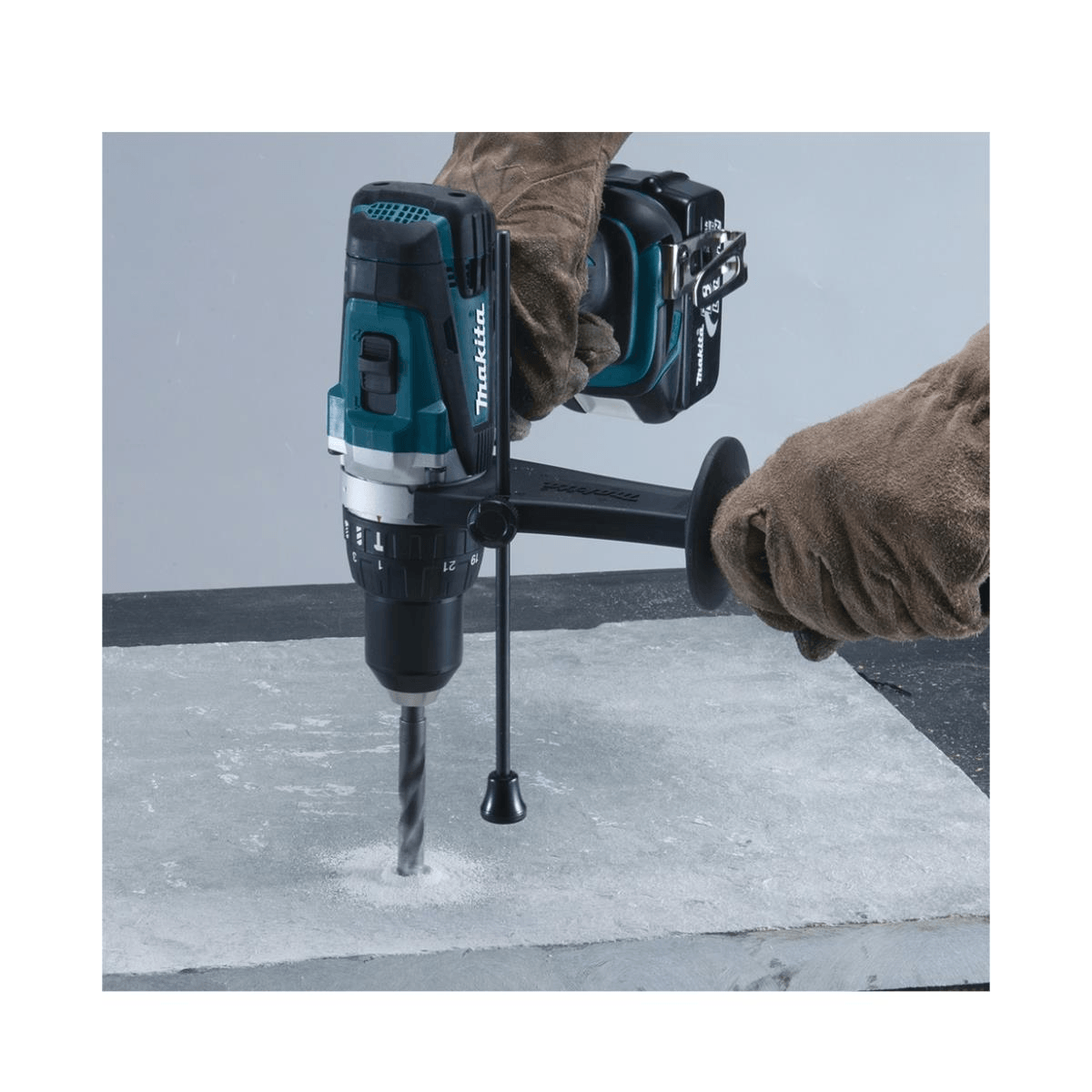 Una persona con guanti da lavoro fora una lastra di cemento con un trapano Makita a batteria.