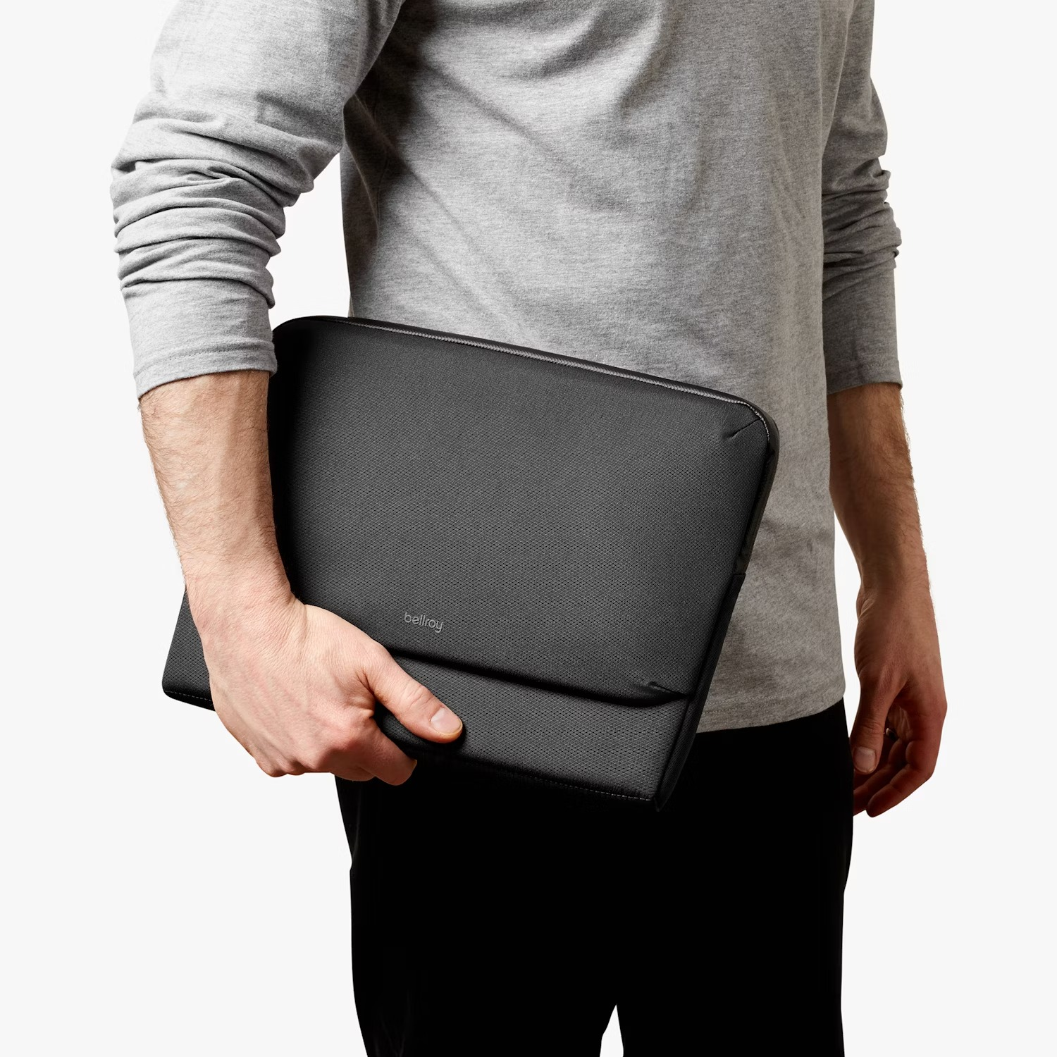Osoba w szarej koszulce z długim rękawem trzyma czarne etui na laptopa z logo Bellroy na białym tle.