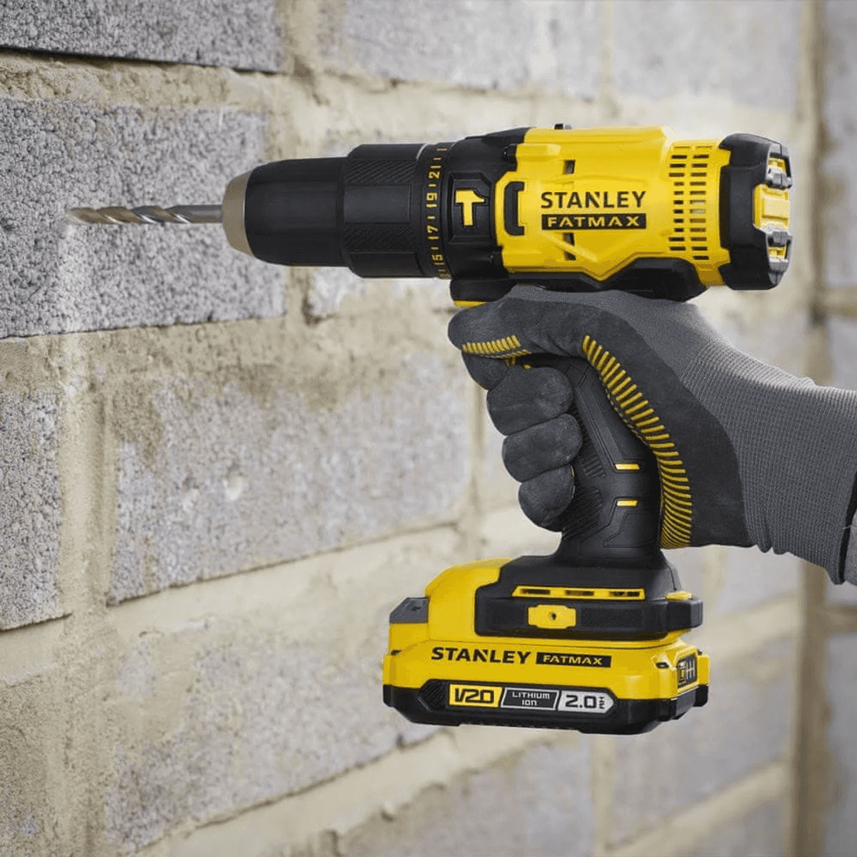 Una mano con guante sostiene un taladro inalámbrico Stanley Fatmax amarillo y negro perforando una pared.