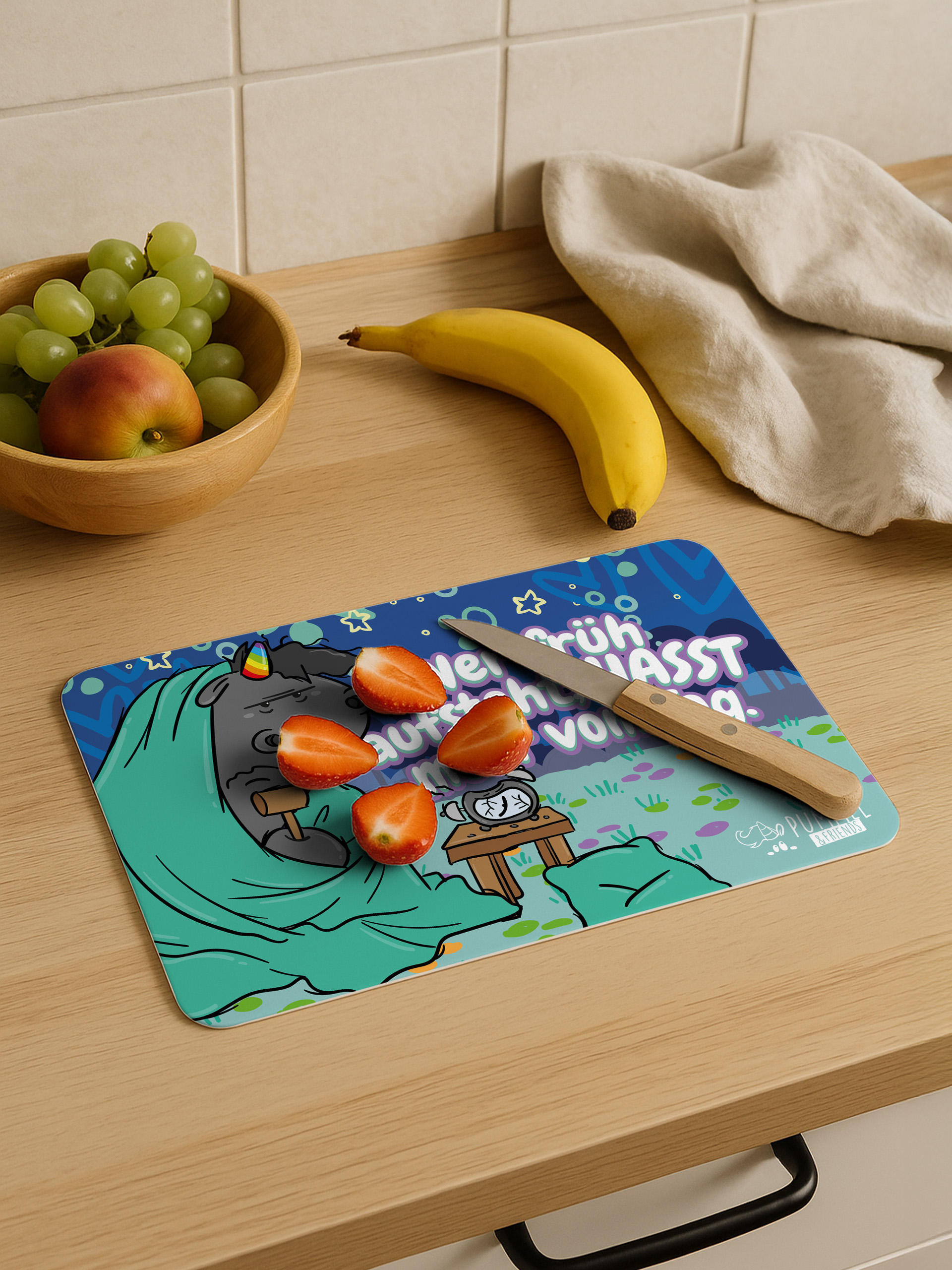 Auf einem Holztisch liegen Erdbeerstücke, ein Messer, eine Banane, eine Obstschale mit Weintrauben und ein Stofftuch.