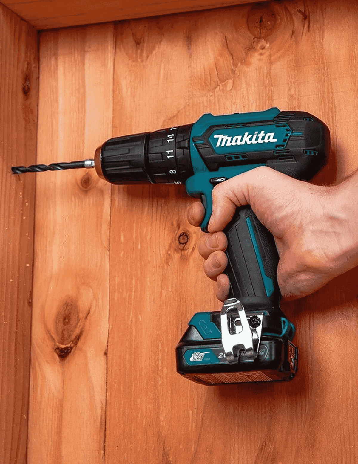 Una mano sostiene un taladro inalámbrico Makita verde azulado y negro contra una pared de madera.