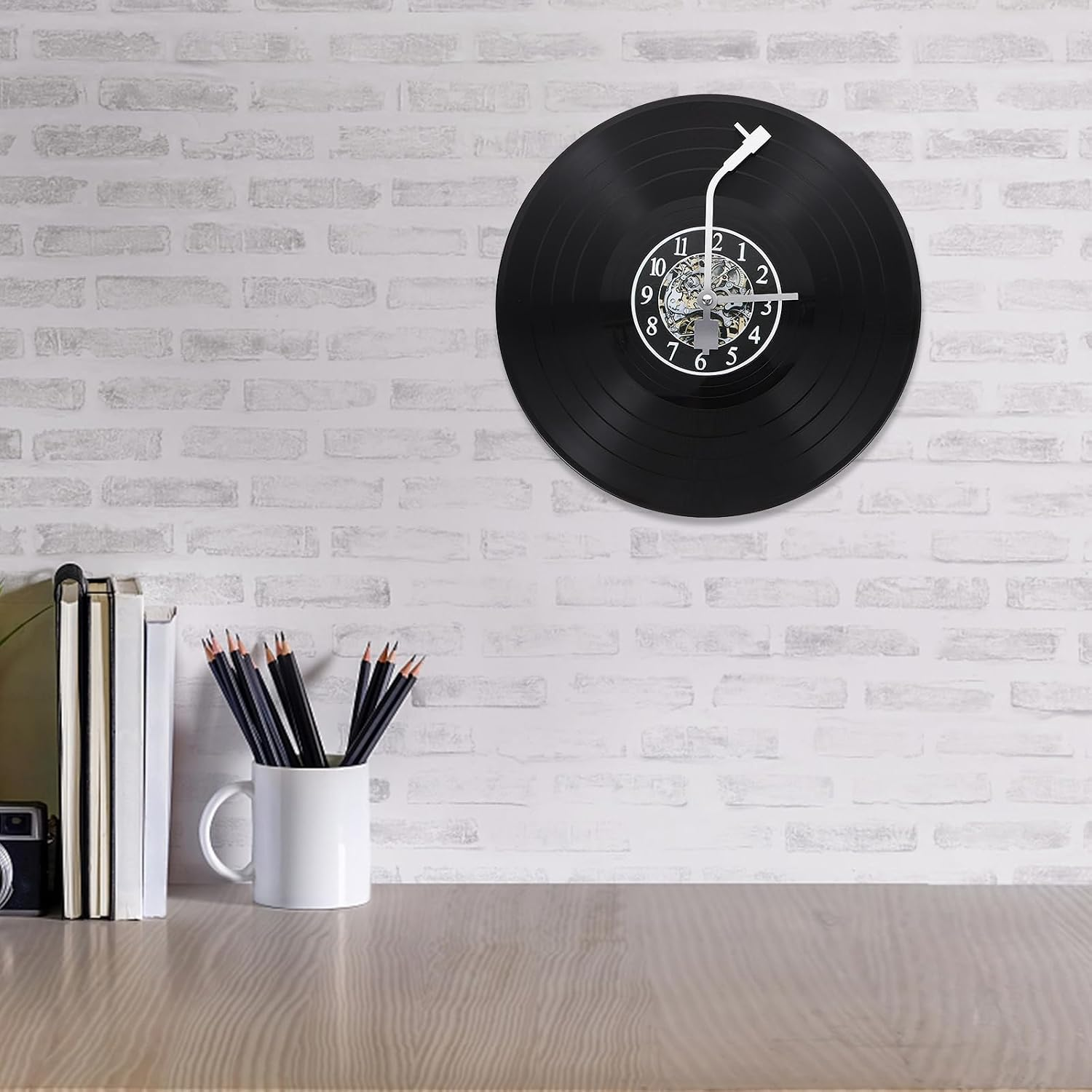 Eine Uhr aus einer schwarzen Schallplatte an einer weißen Backsteinwand über einem Holztisch mit Büchern und Bleistiften.