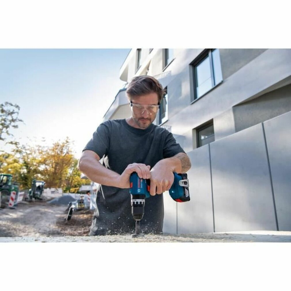 Un homme portant des lunettes de protection utilise une perceuse bleue et noire près d'un bâtiment.
