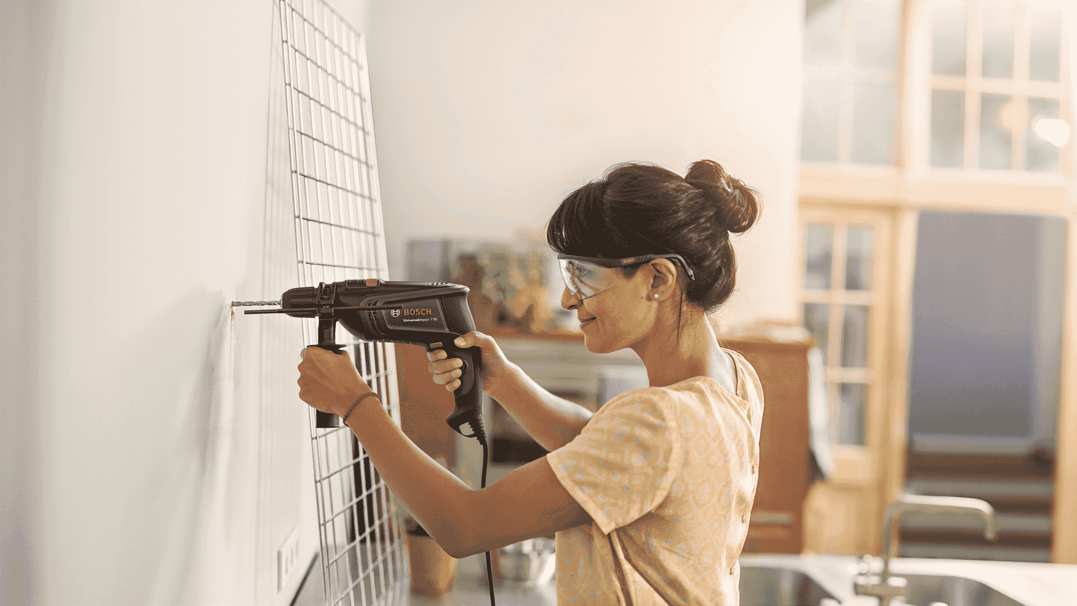 Una donna con occhiali protettivi usa un trapano Bosch su una parete con griglia.