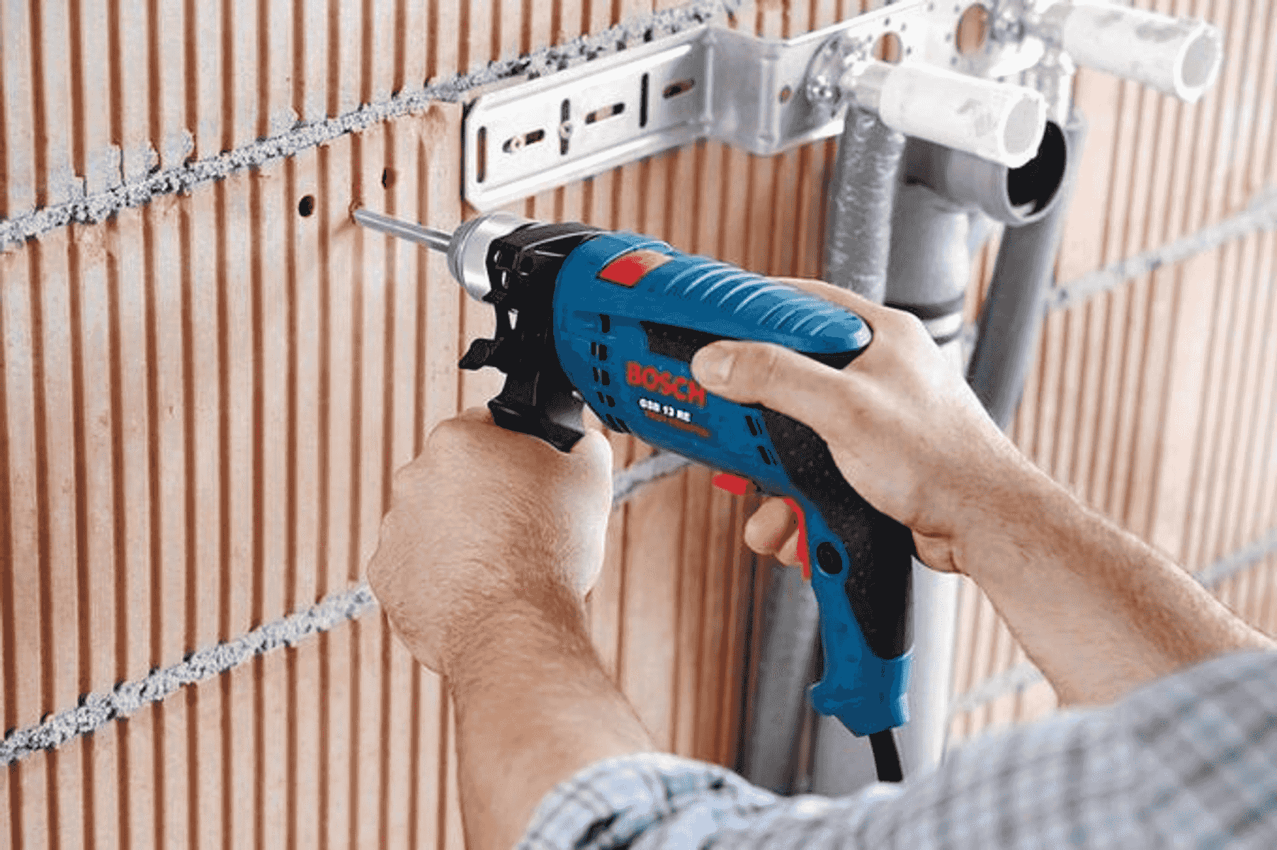 Bir kişi tuğla duvara delik açmak için mavi bir Bosch matkap kullanıyor.