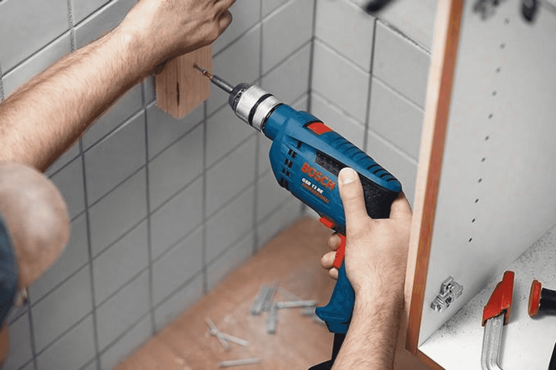 Bir kişi fayanslı bir duvara karşı ahşap bir blokta mavi bir Bosch matkap kullanıyor.