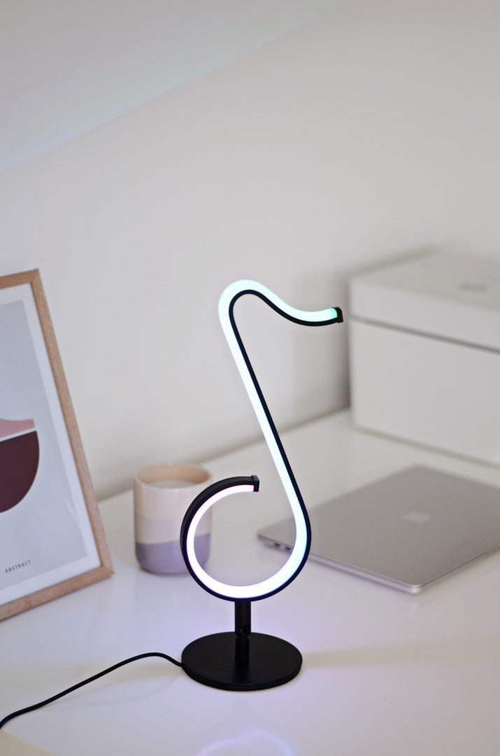 Una lampada da scrivania nera con una striscia luminosa bianca a forma di nota musicale su un tavolo bianco.