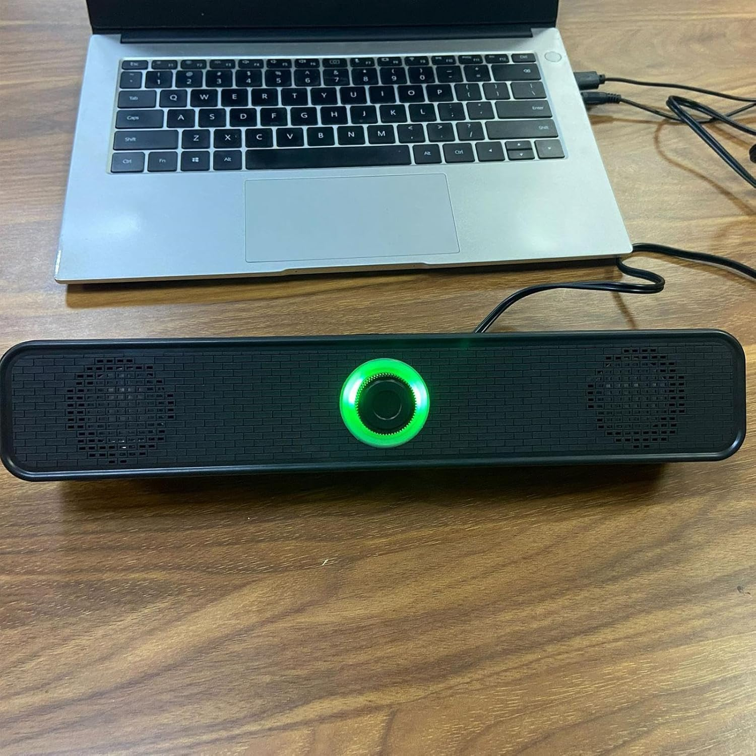 Eine lange schwarze Soundbar mit grün leuchtendem Drehregler liegt vor einem Laptop auf Holztisch.