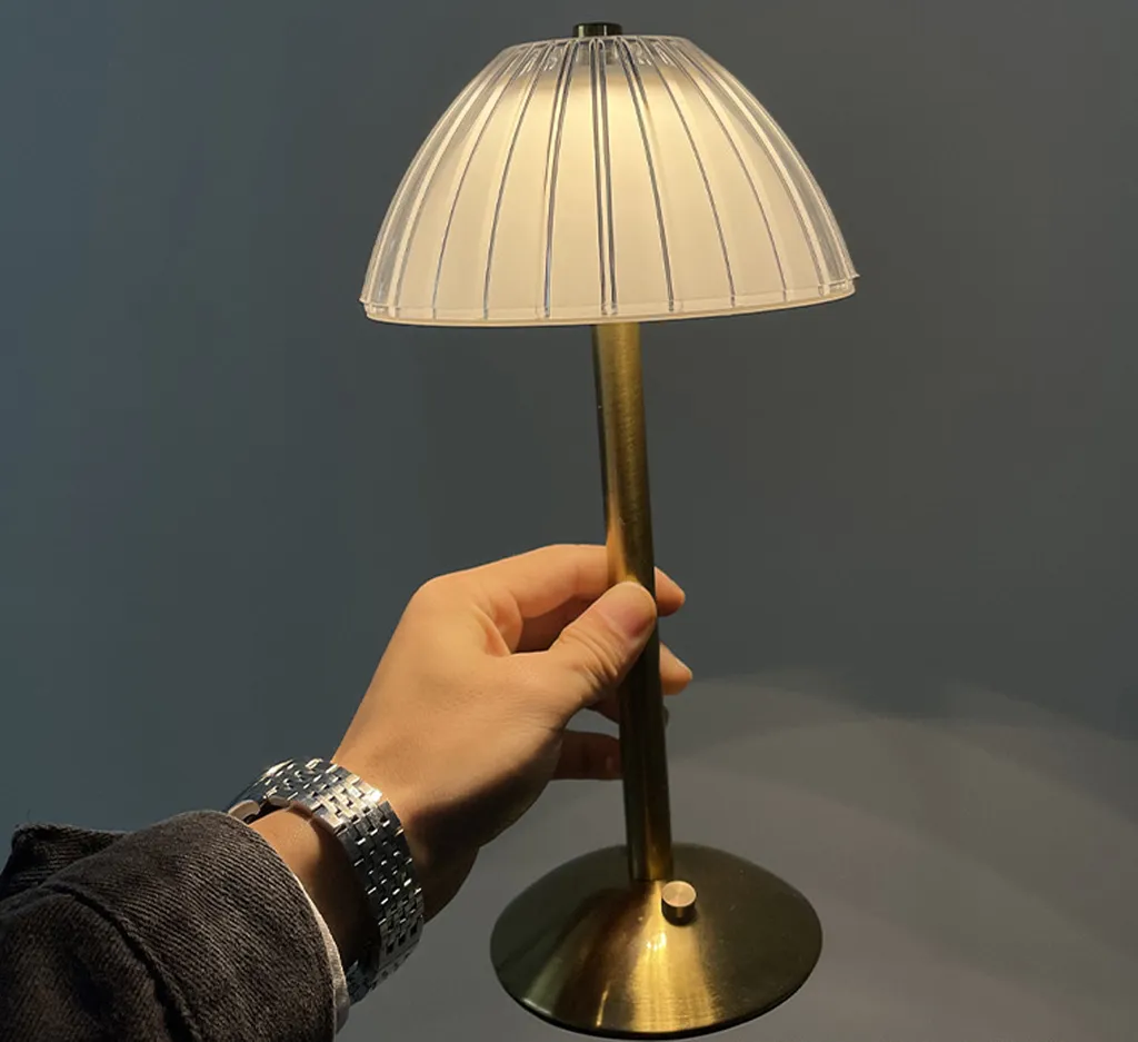 Eine Hand hält eine kleine, moderne Tischlampe mit goldenem Ständer und weißem Schirm vor einem dunklen Hintergrund.