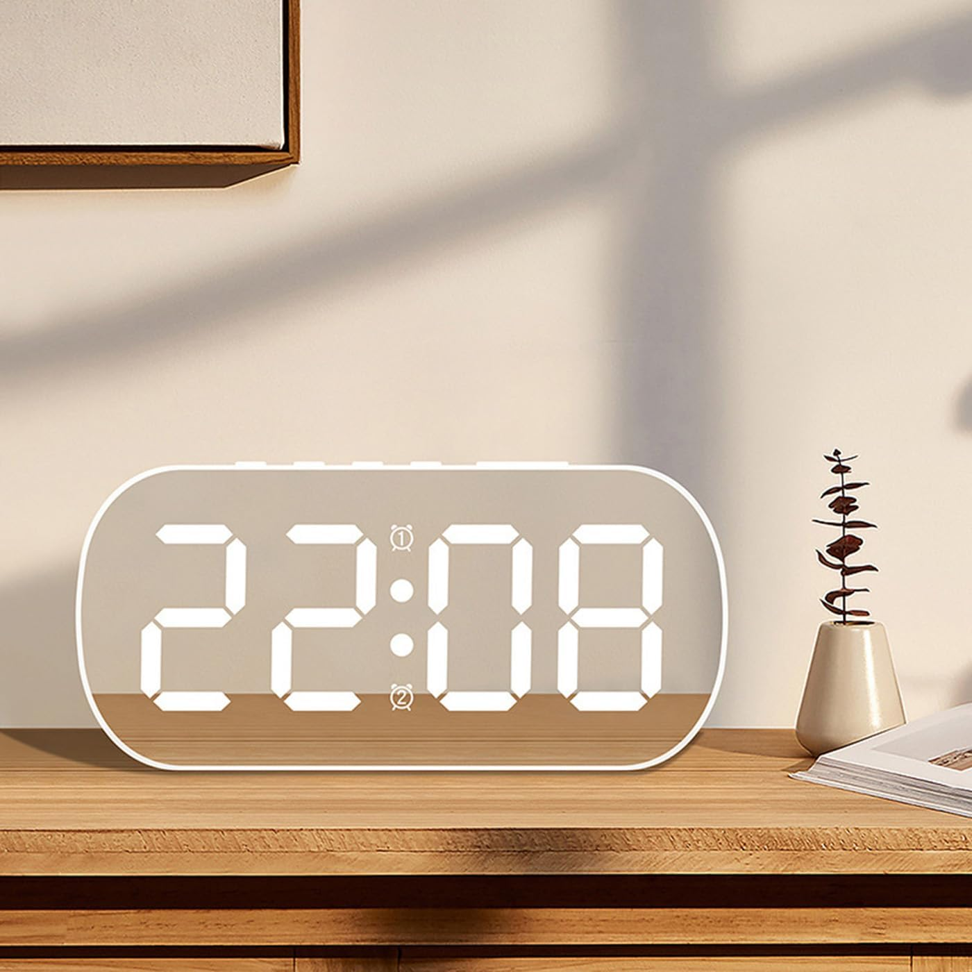 Ein digitaler Wecker auf einem Holztisch zeigt 22:08, neben einer kleinen Vase mit Blumen und einem Buch.