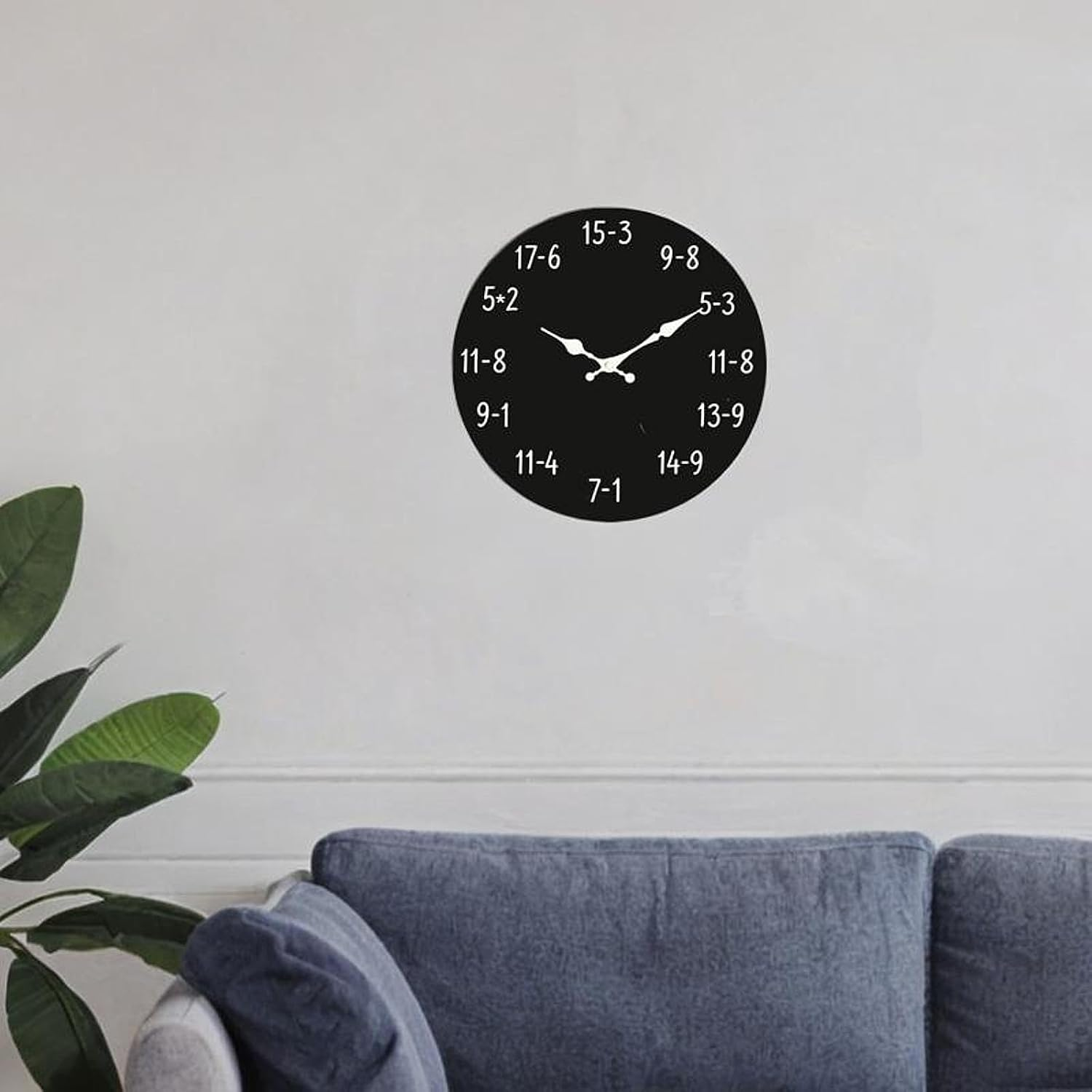 Eine schwarze Mathe-Wanduhr über einem blauen Sofa, mit einer grünen Pflanze im linken Vordergrund.