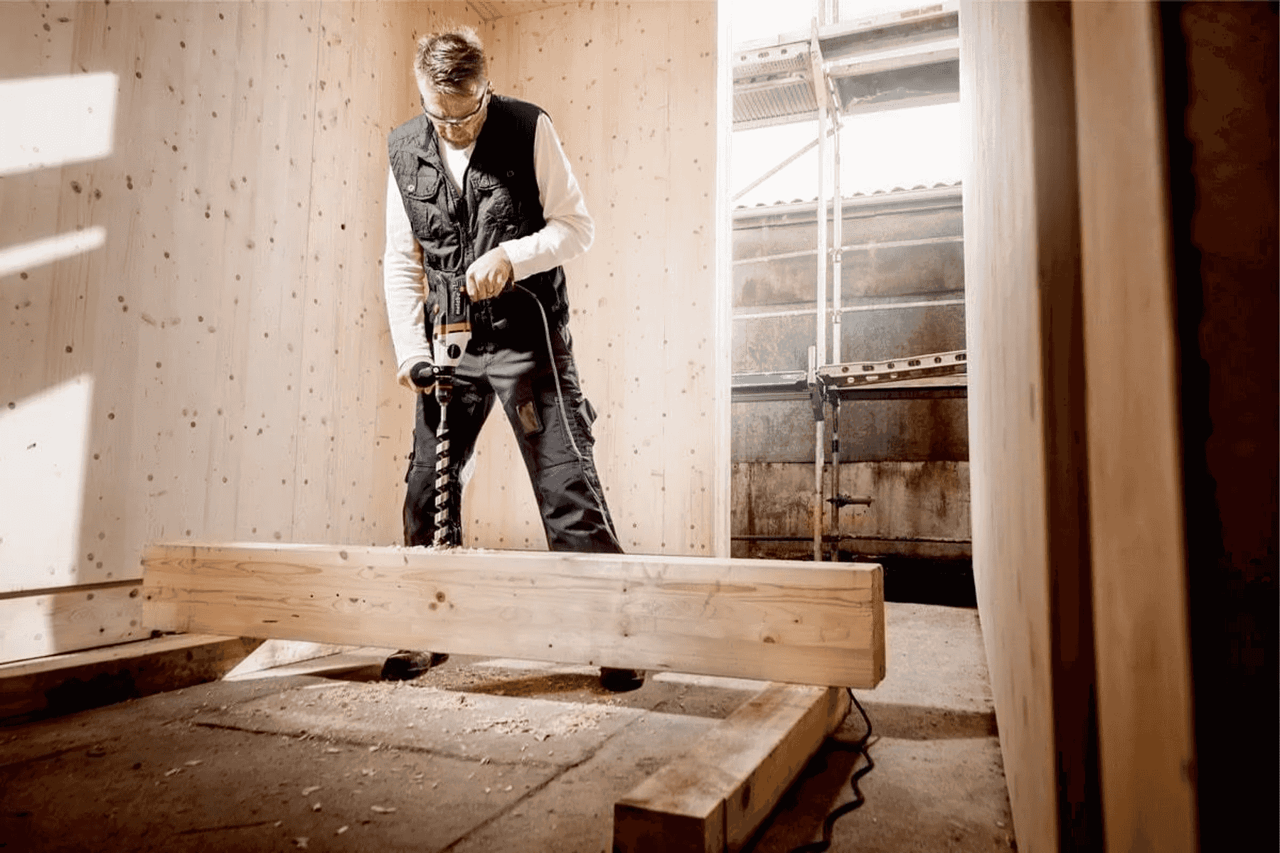 Un hombre con gafas de seguridad y chaleco taladra una viga de madera en el interior.