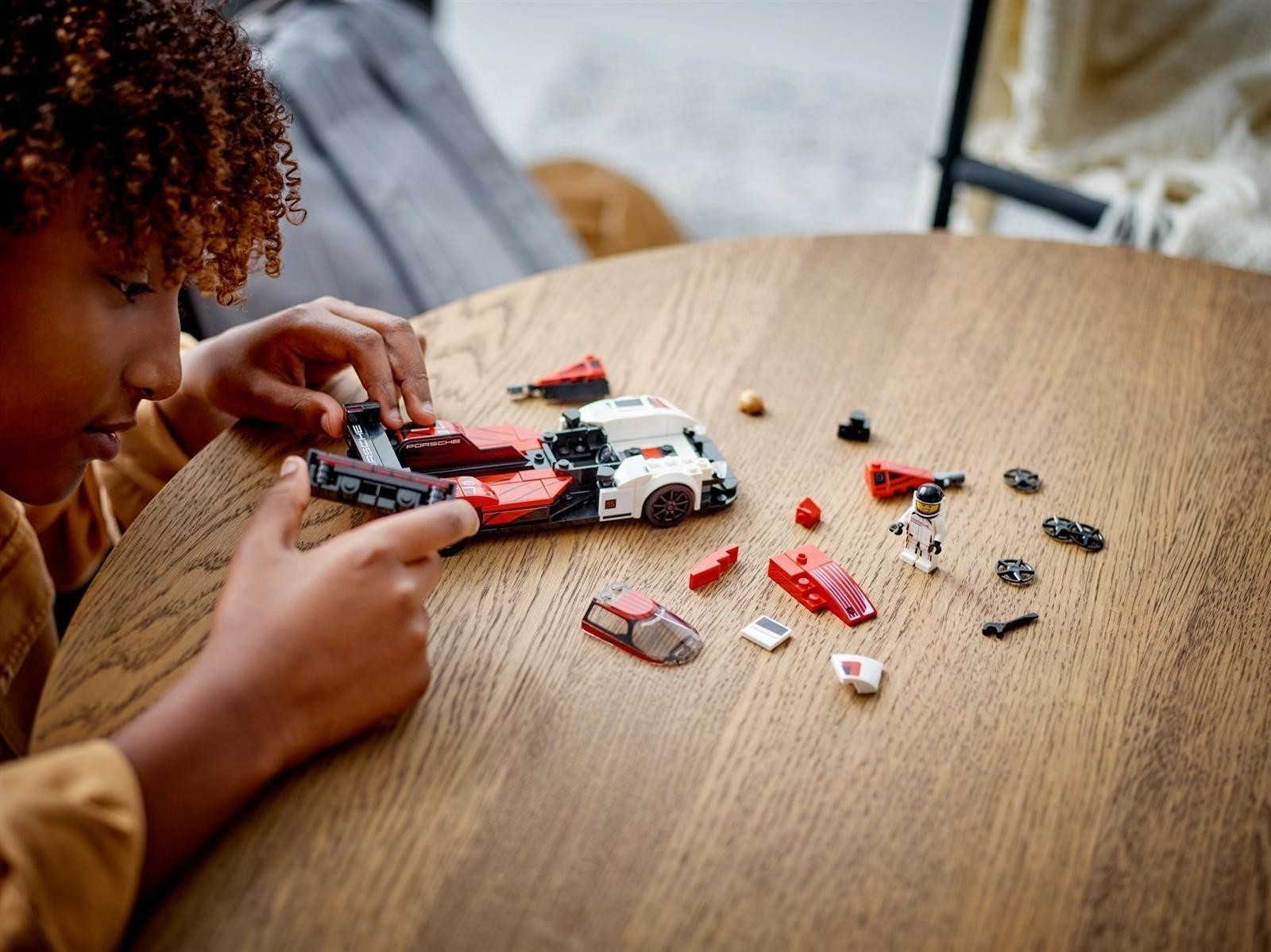 Ein Kind baut ein rot-weißes Lego-Rennauto auf einem Holztisch, umgeben von losen Teilen und einer Minifigur.