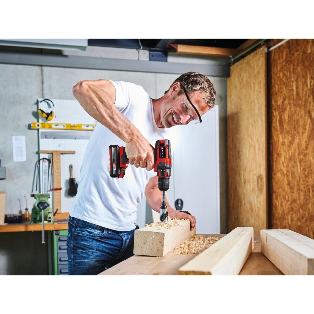 Un hombre con camiseta blanca perfora una viga de madera en un taller.