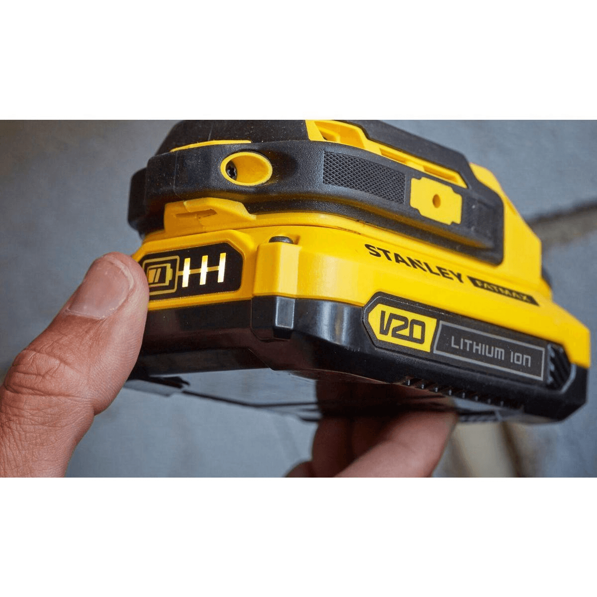 Una mano sostiene una batería de litio Stanley V20 amarilla y negra con barras indicadoras de carga iluminadas.