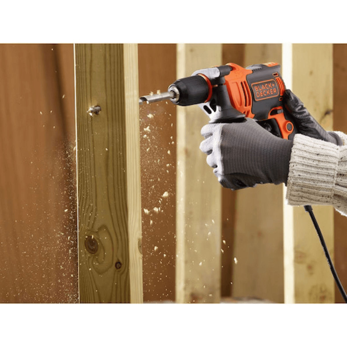 Persona perforando una tabla de madera con un taladro naranja y negro, virutas volando.