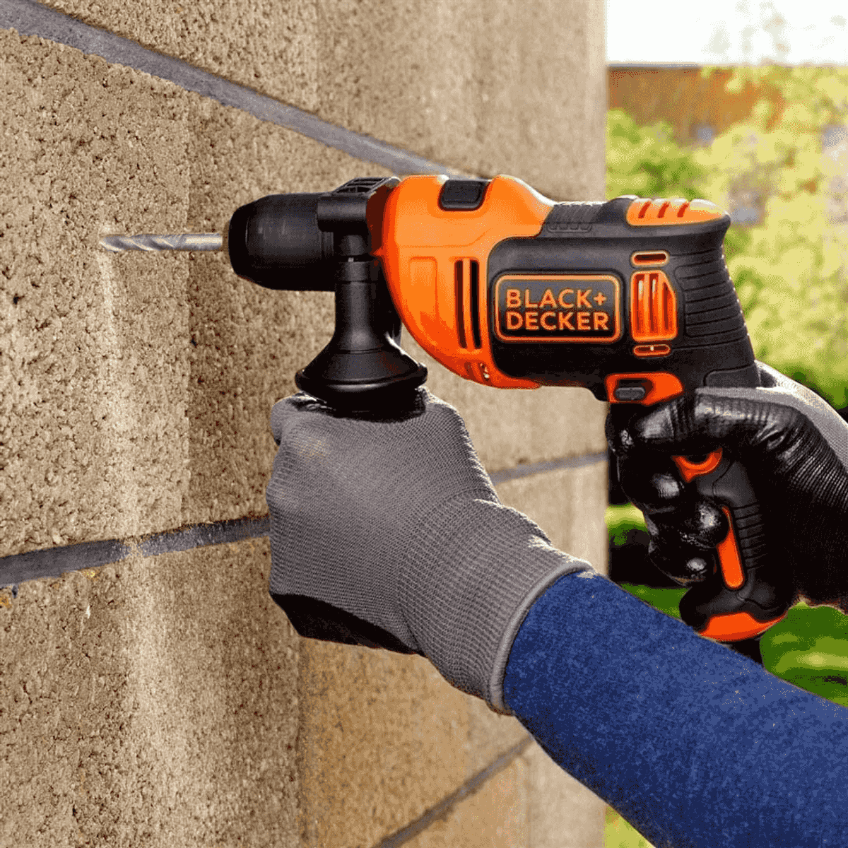 Una mano enguantada sostiene un taladro Black+Decker naranja y negro perforando una pared de bloques de hormigón.