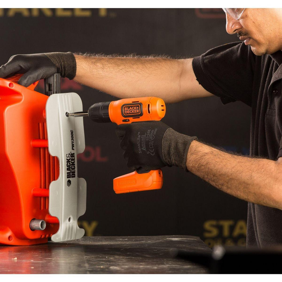 Un hombre con guantes usa un taladro inalámbrico naranja Black+Decker en un dispositivo naranja.